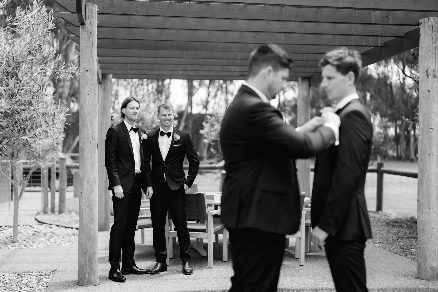 Two people in black tuxedos helping each other with bow ties while others watch in a black and white outdoor setting.