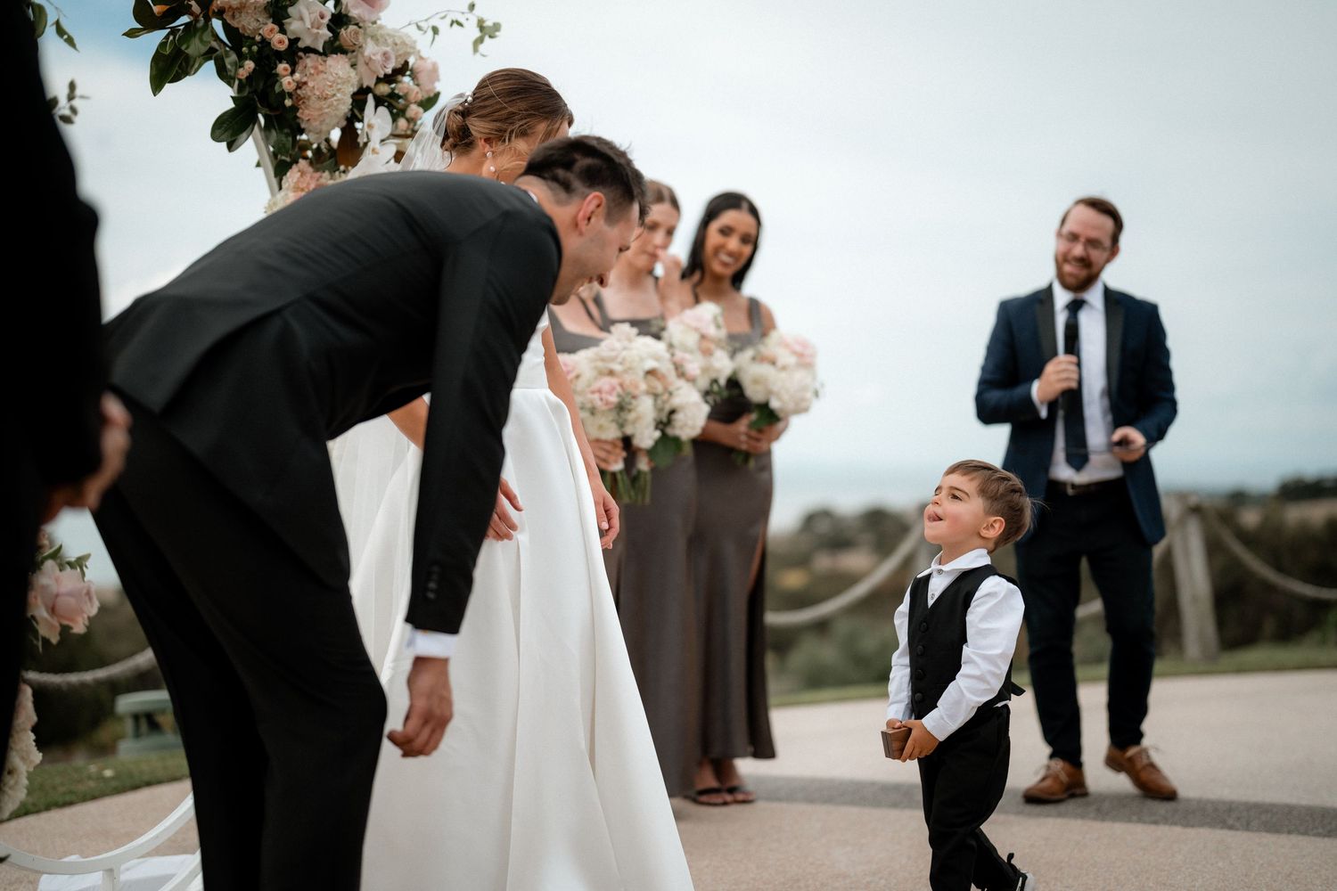 A young ring bearer in a vest shares a sweet moment with wedding party members on a path during an outdoor ceremony.