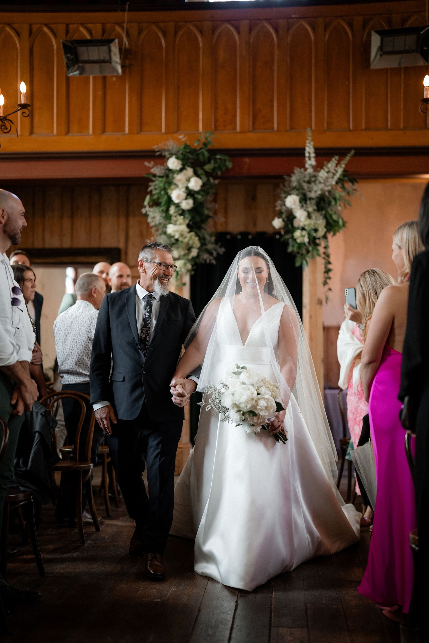 Elegant wedding ceremony in a rustic wooden venue with floral arrangements and guests in formal attire.
