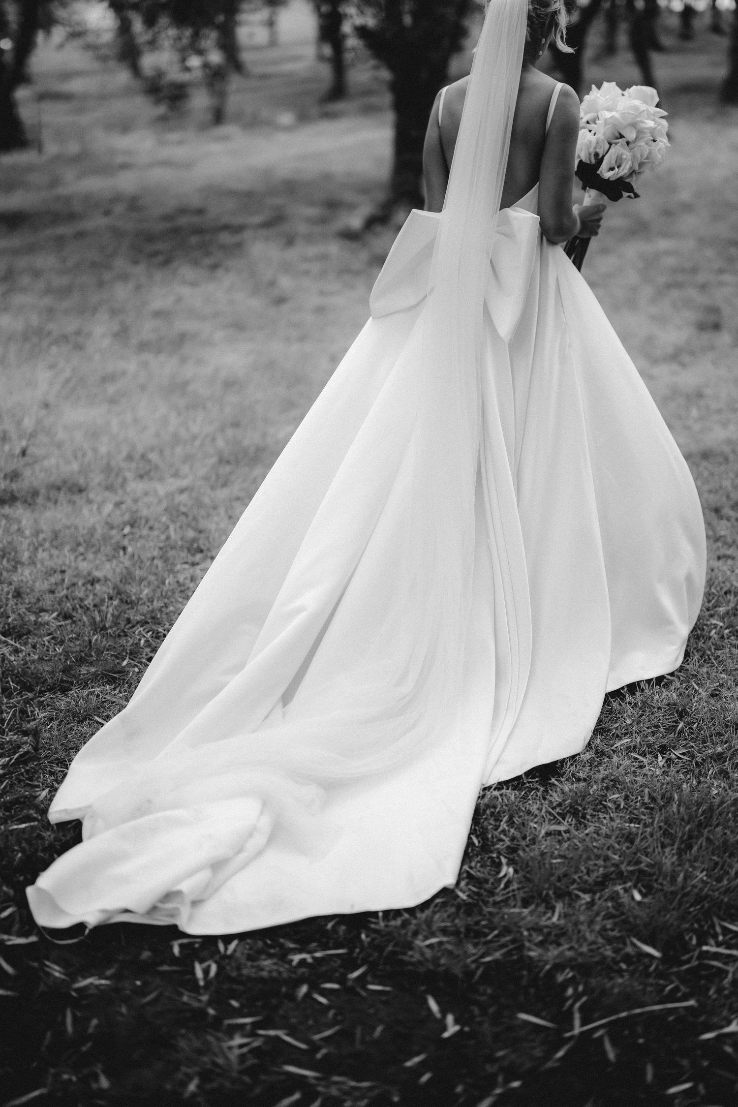 Black and white photo of a bride's flowing wedding dress train trailing behind her as she walks on grass holding flowers.