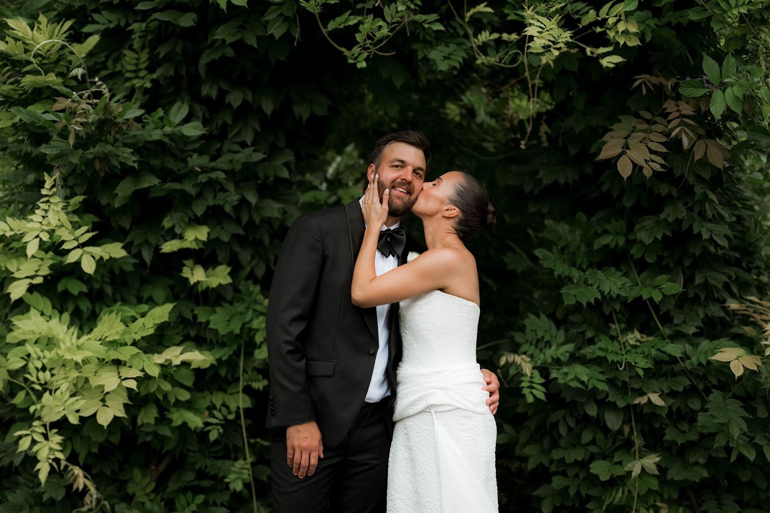 Bride and groom share a tender kiss surrounded by lush green foliage on their wedding day.