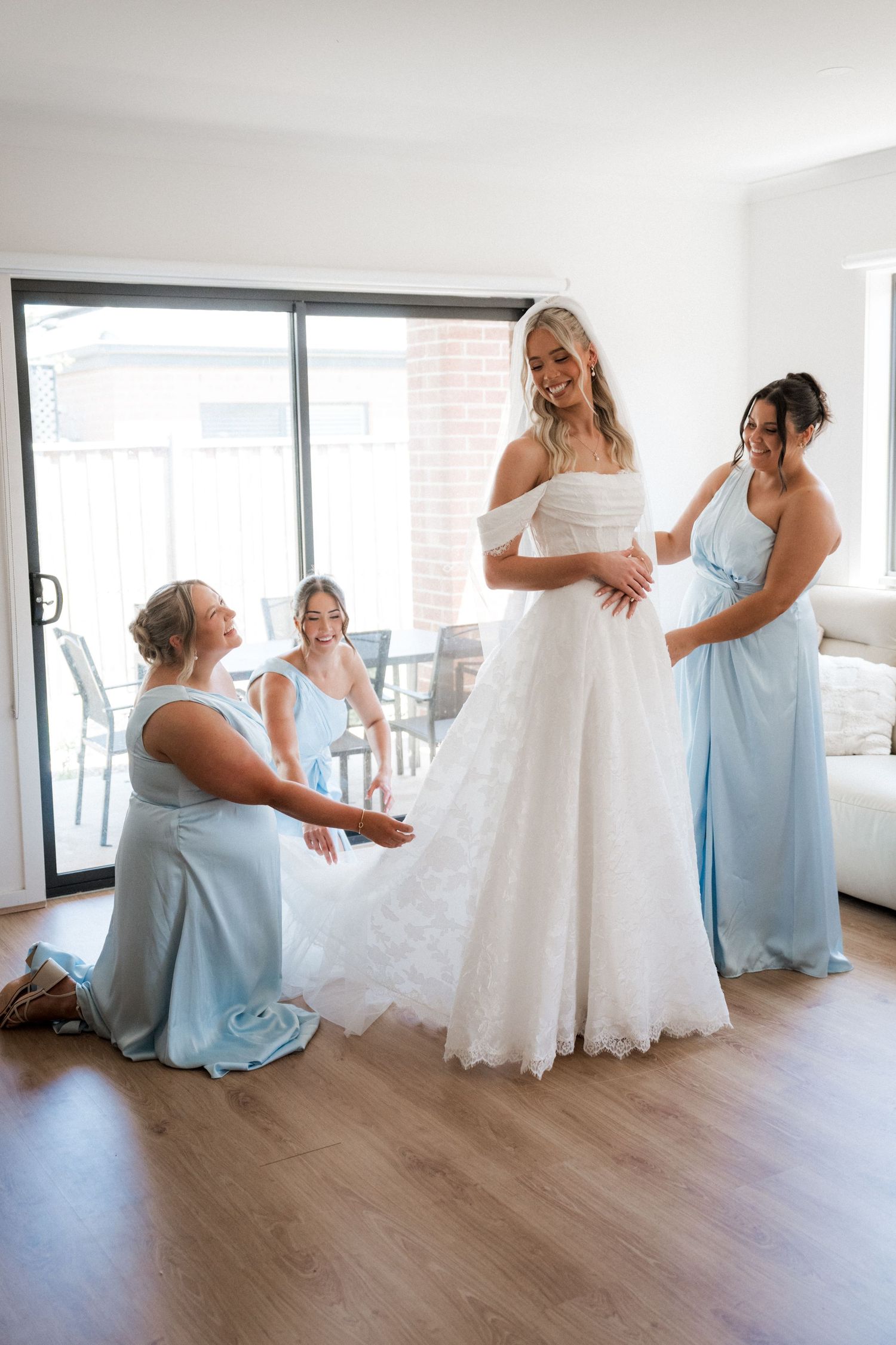 Bride in off-shoulder lace gown assisted by three bridesmaids in light blue dresses while getting ready.