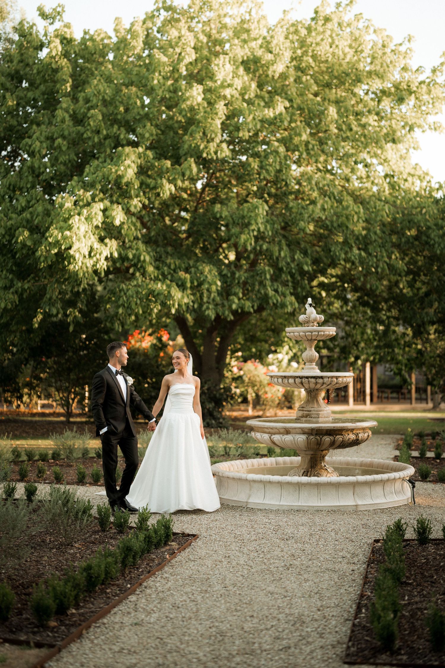 Romantic wedding portrait by ornate garden fountain surrounded by lush greenery.