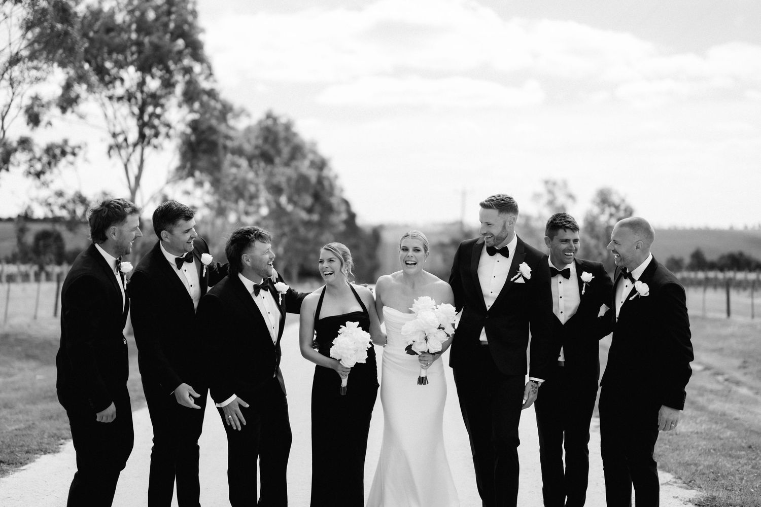 Wedding party in formal black attire stands in line against rustic outdoor backdrop.
