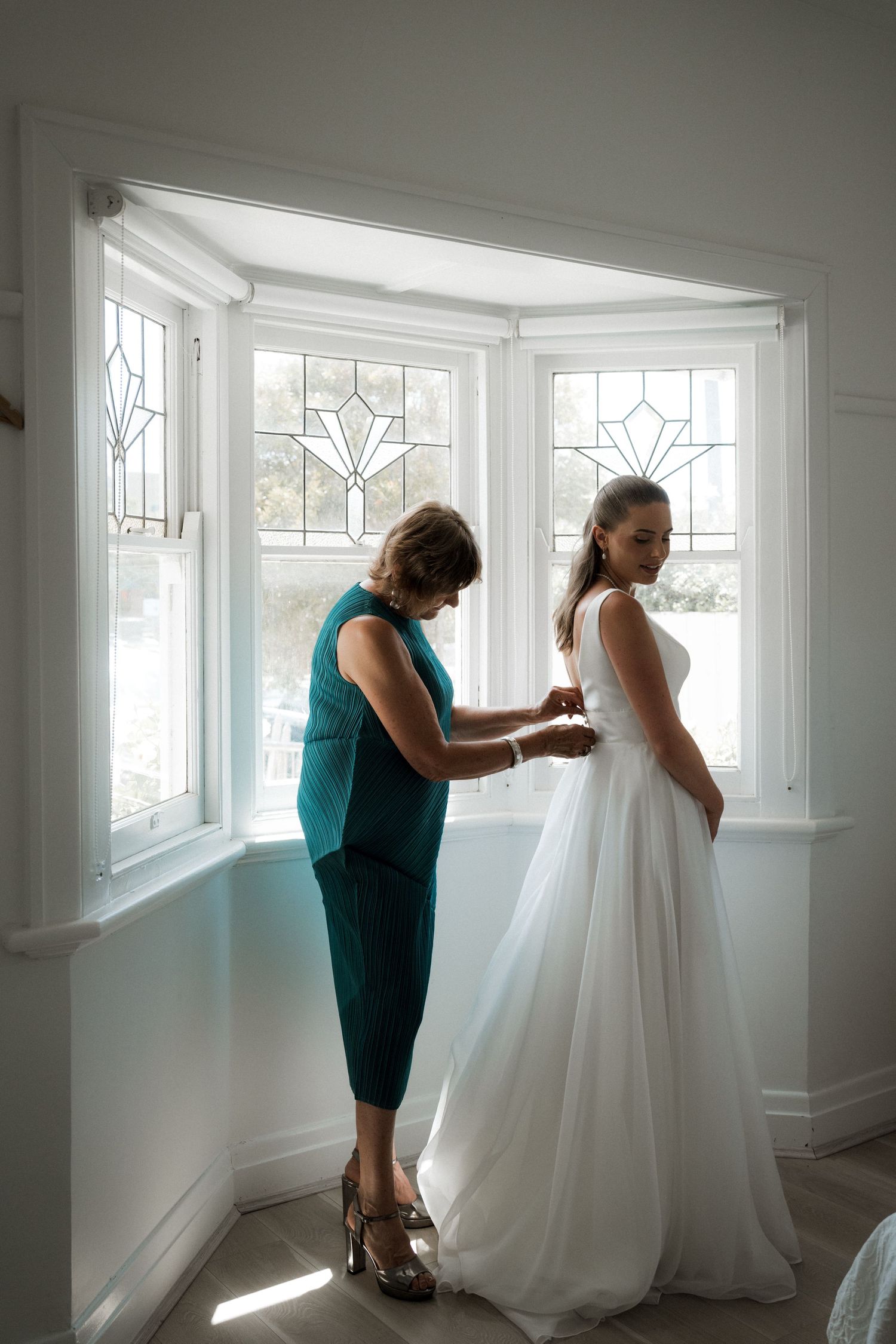 A bridesmaid in teal helps adjust a white wedding dress near sunlit windows.