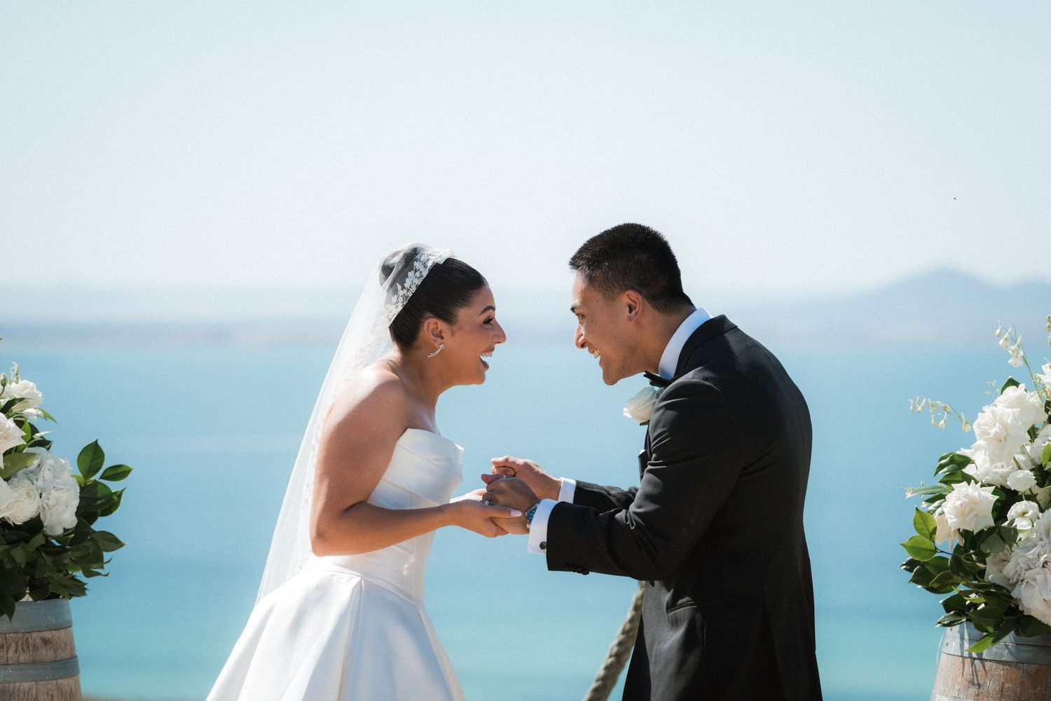 Couple shares intimate moments during their oceanfront wedding ceremony with white floral decorations and blue waters.