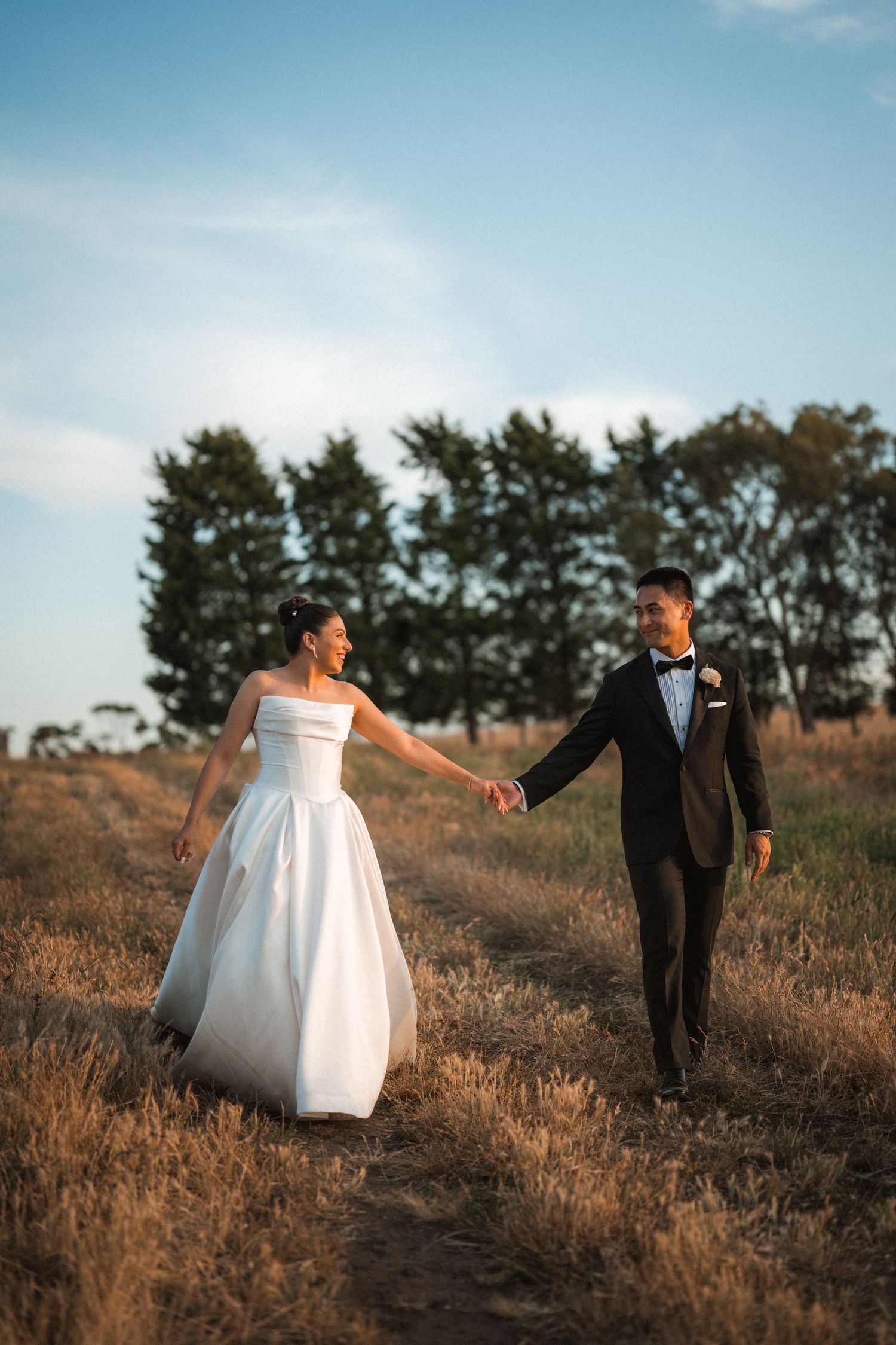 Newlyweds walk hand in hand through a golden field at sunset.