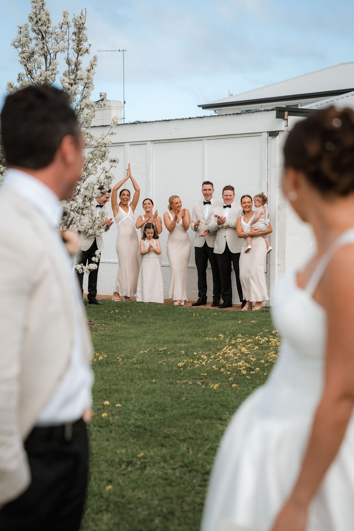 Wedding party in white and neutral attire poses together on a grassy lawn.