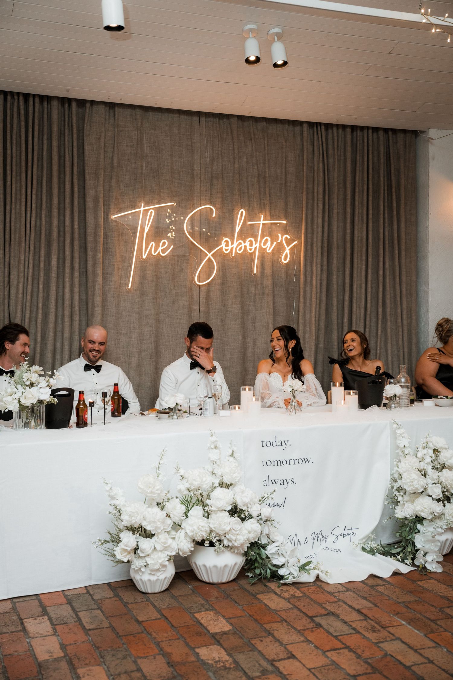 Wedding reception head table decorated with white flowers and neon sign backdrop.