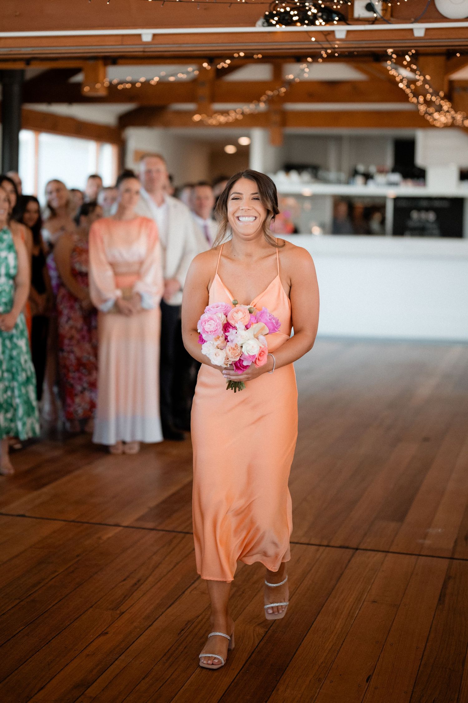 Wedding party member in peach dress walking across wooden floor with pink bouquet.