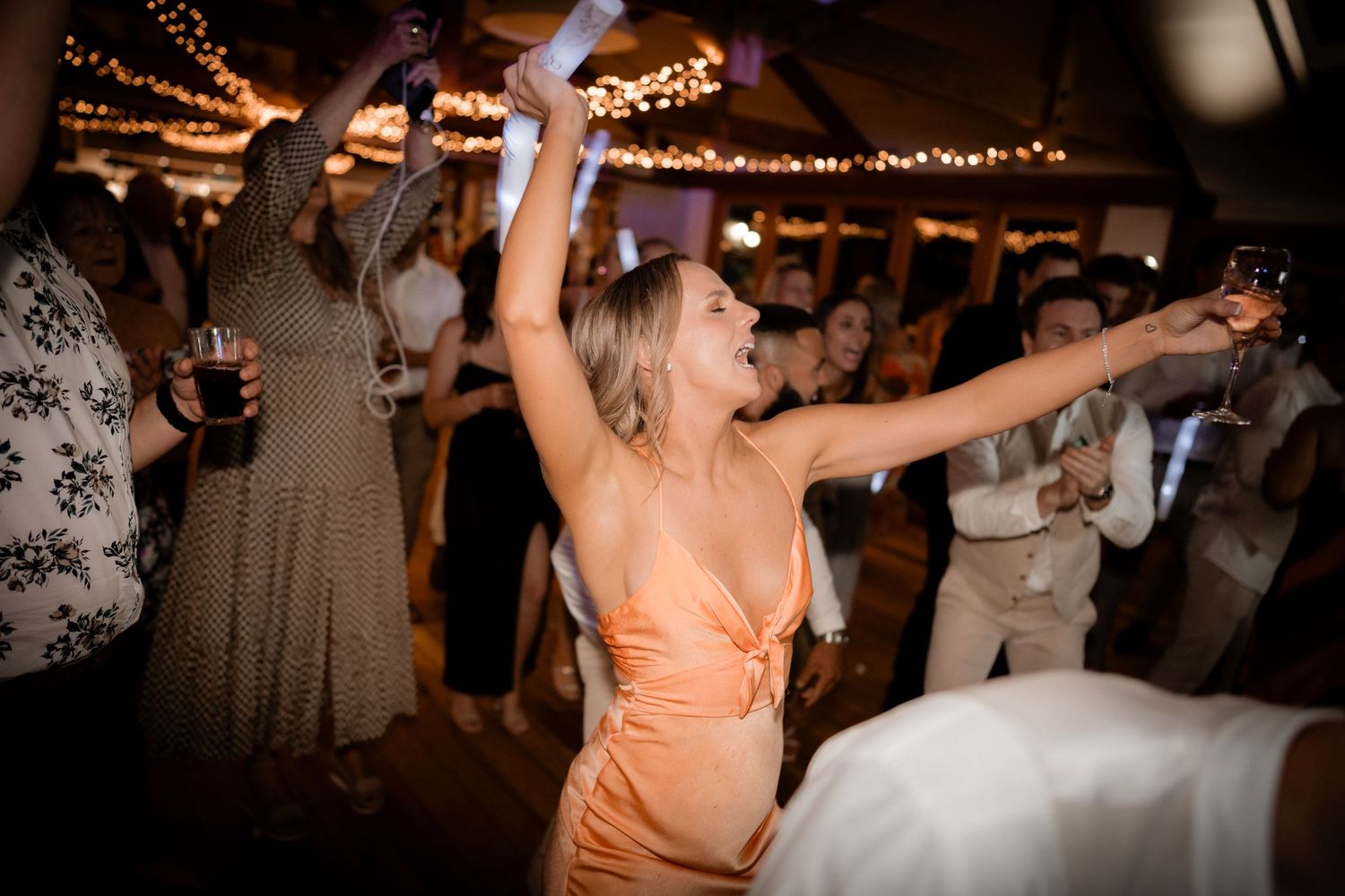 People dancing and celebrating at a wedding reception under glowing string lights and wooden beams.