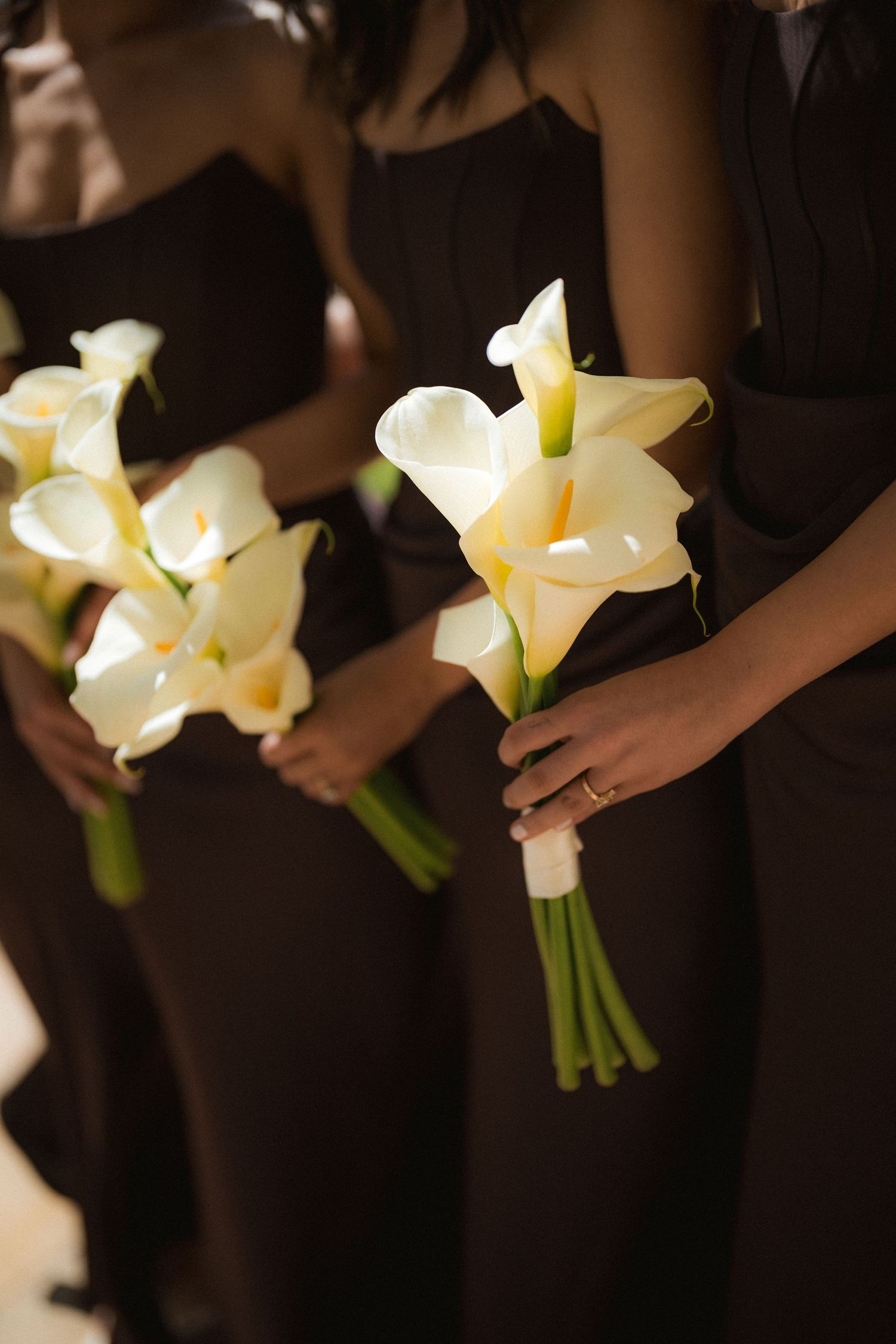Bridesmaids in dark brown dresses hold bouquets of white calla lilies with green stems at a wedding ceremony.