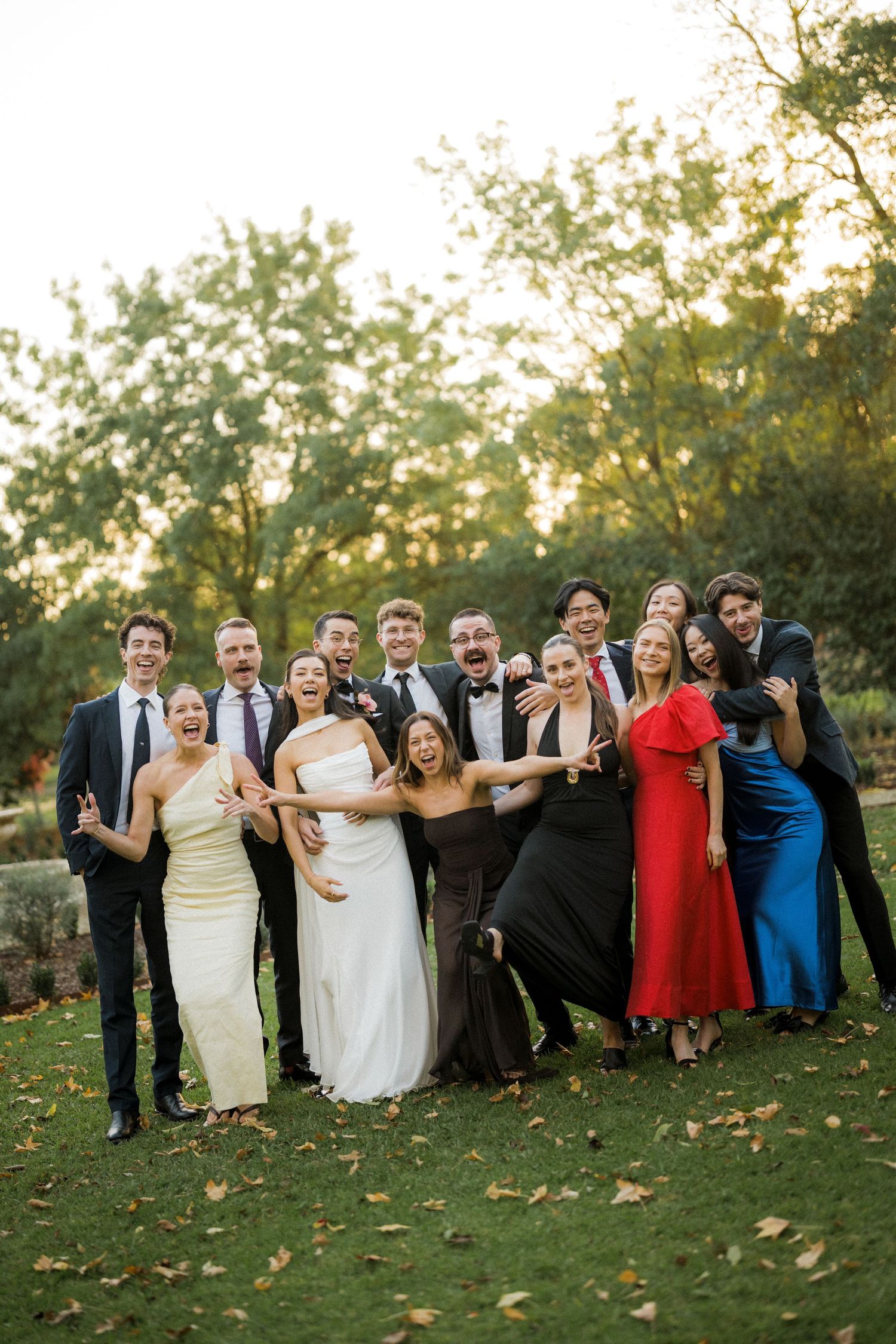 Joyful wedding party group photo with bride, groom and guests celebrating on garden lawn.