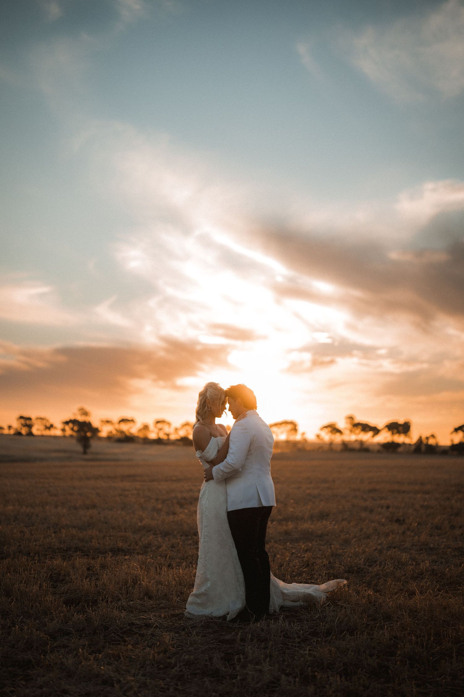 Bride and groom embracing in open field with dramatic golden sunset sky behind them.