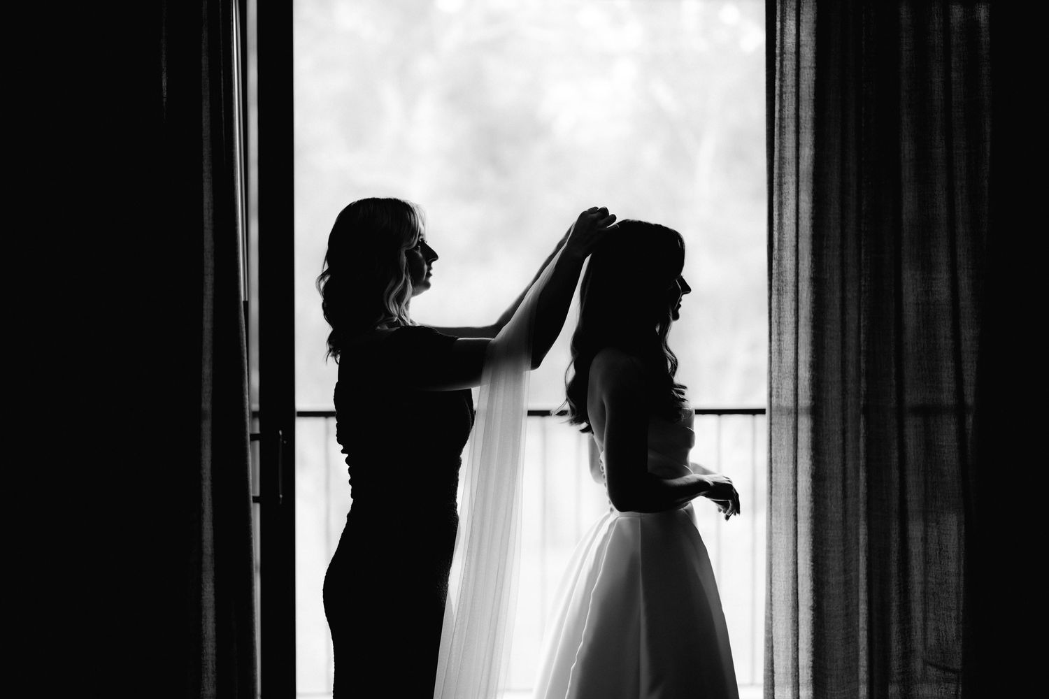 A silhouette shows a wedding preparation moment as the veil is adjusted by the window.