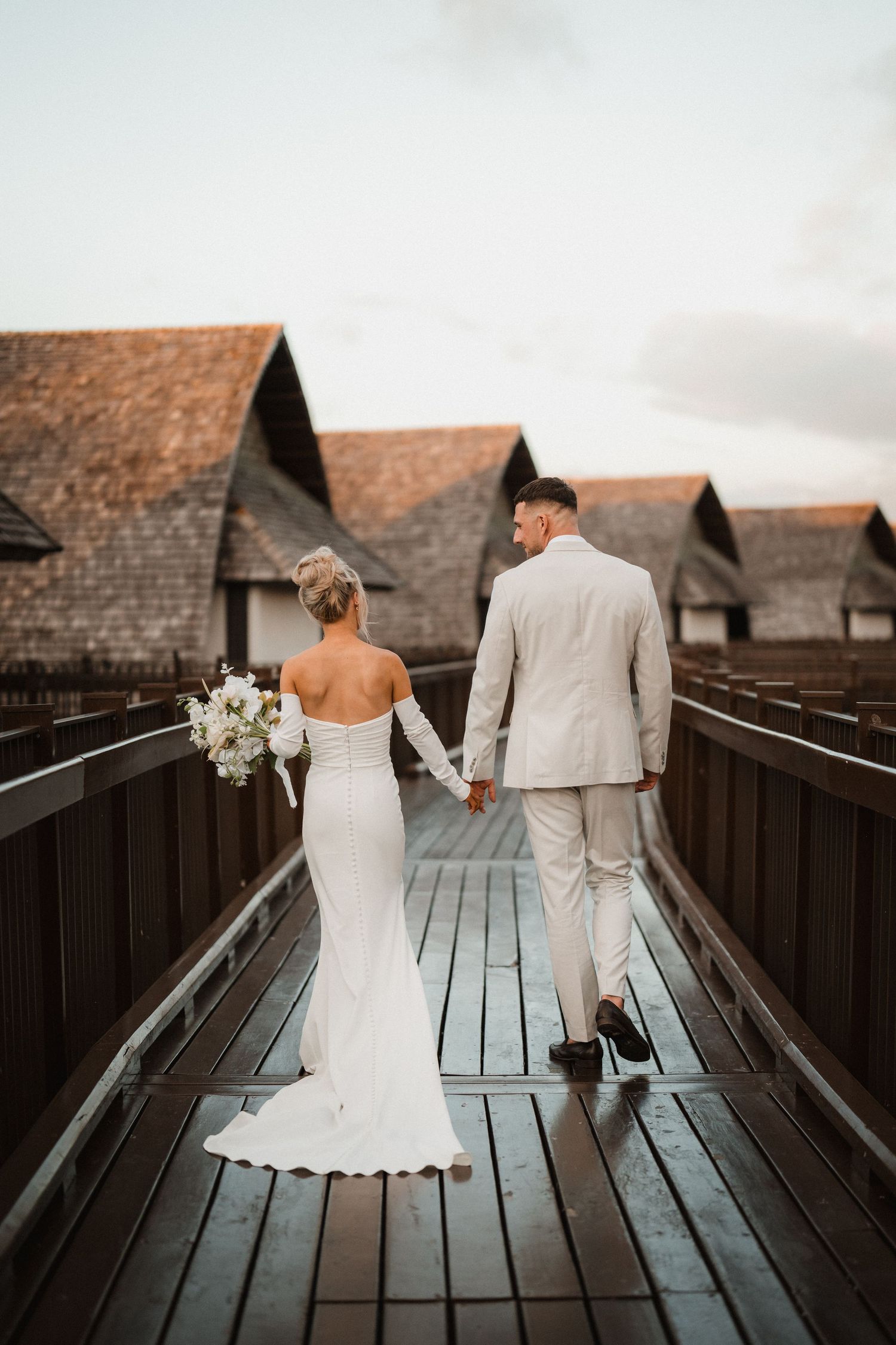 A couple in wedding attire walk along a wooden boardwalk with rustic cabins in the background at sunset.