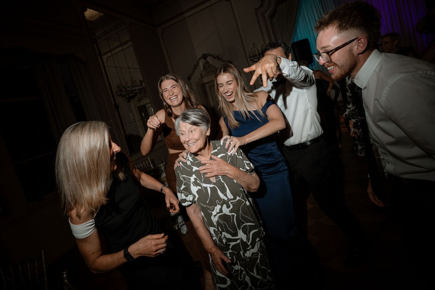 Group of guests dancing and celebrating energetically at a dimly lit wedding reception.