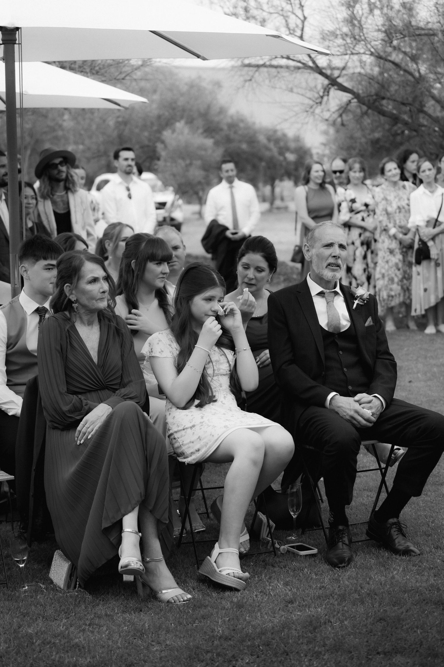 Emotional guests watching a wedding ceremony outdoors under towering trees with varied seating arrangements.