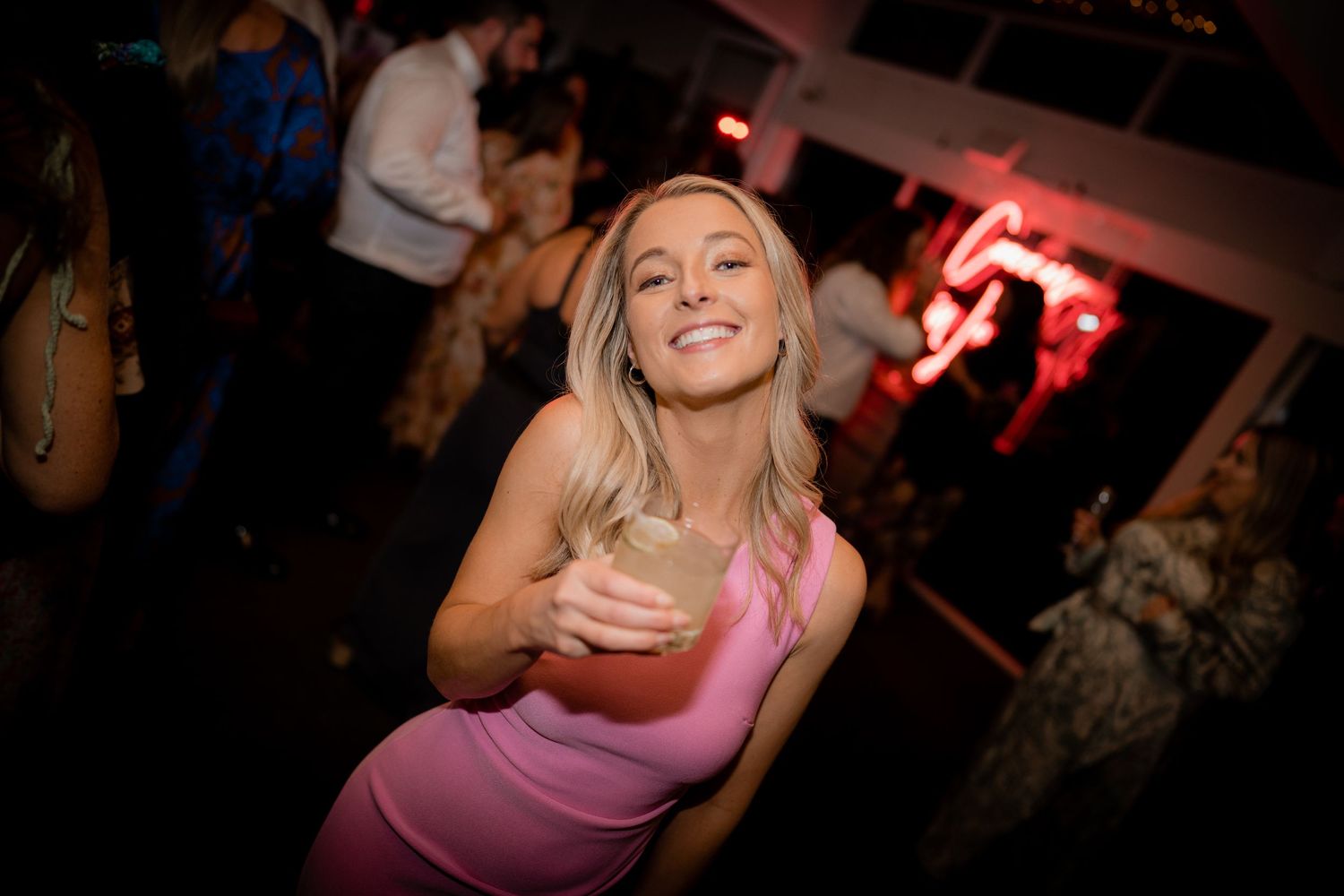 Person in pink dress smiling and dancing at nightclub with drink in hand and party lights in background.