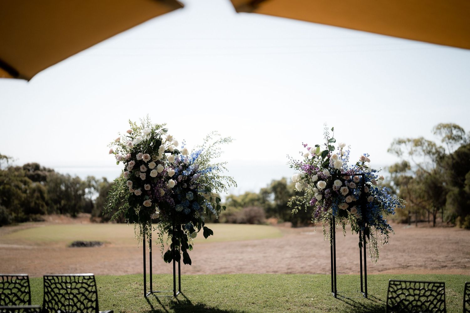 Two metal chairs and floral arrangements frame an outdoor wedding ceremony space against a natural landscape backdrop.