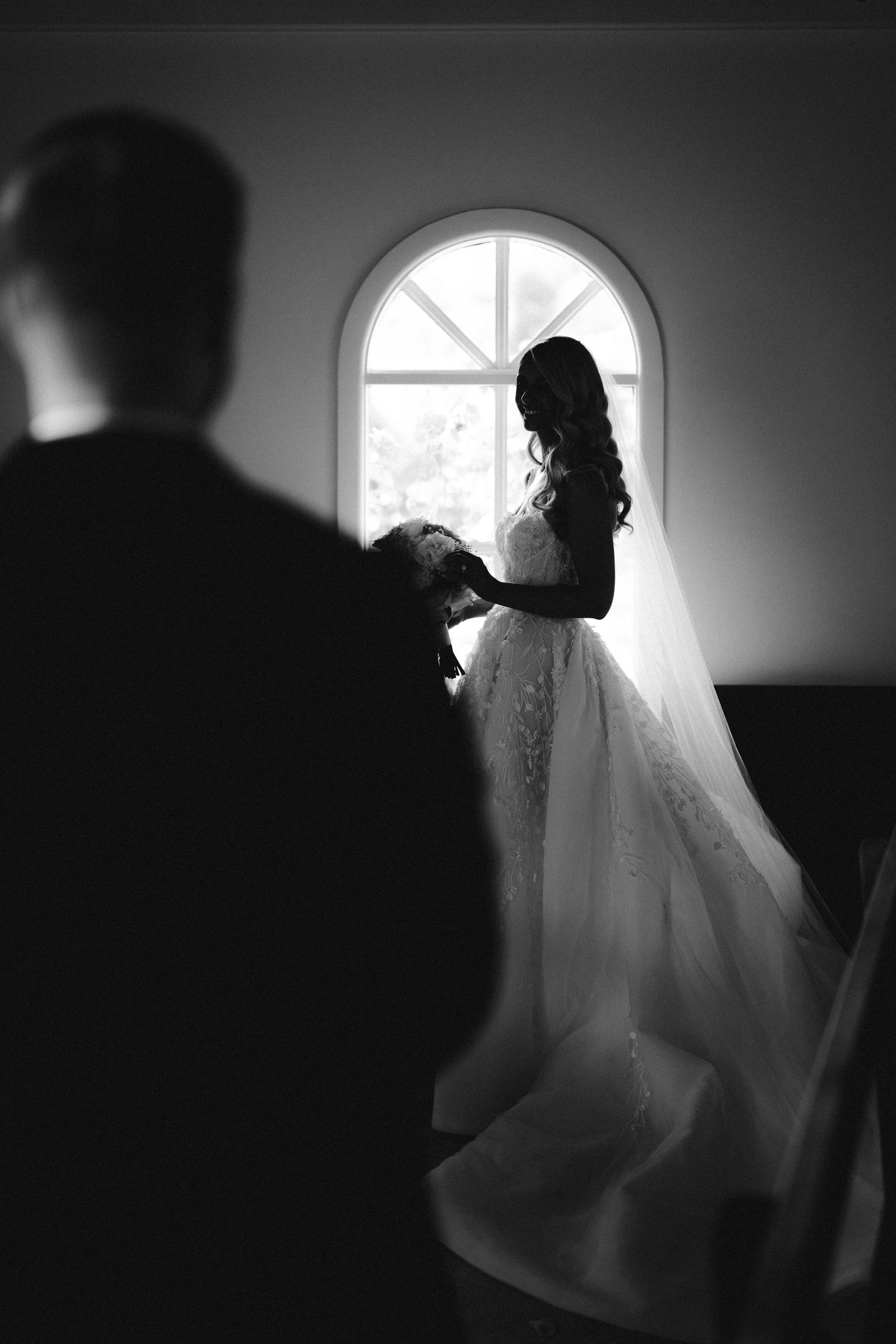 Dramatic silhouette of a bride in front of an arched window in black and white.