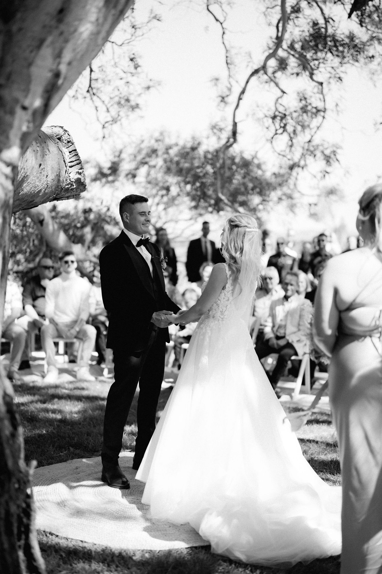 Bride and groom exchange vows during outdoor garden wedding ceremony with guests seated beneath shady trees in black and white.
