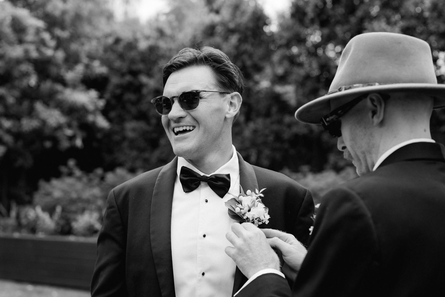 A person in a black tuxedo and bowtie smiles joyfully while someone in a hat adjusts their boutonniere outdoors.