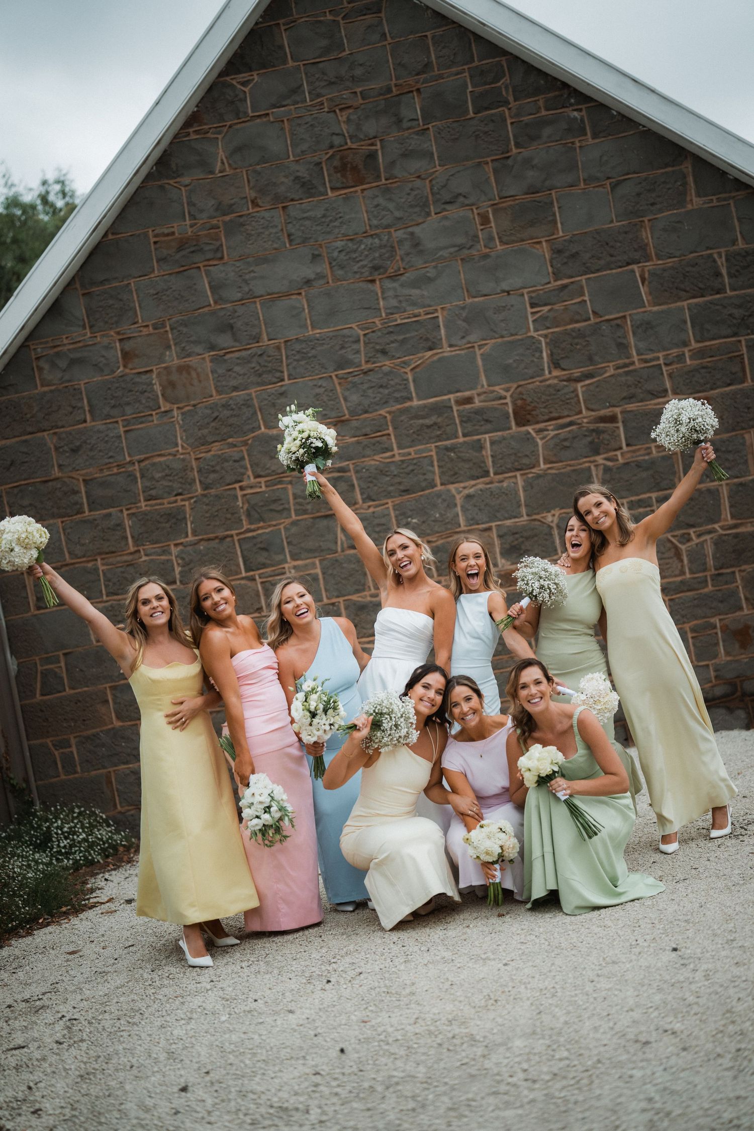 Bridesmaids in pastel dresses pose with bouquets against stone wall.