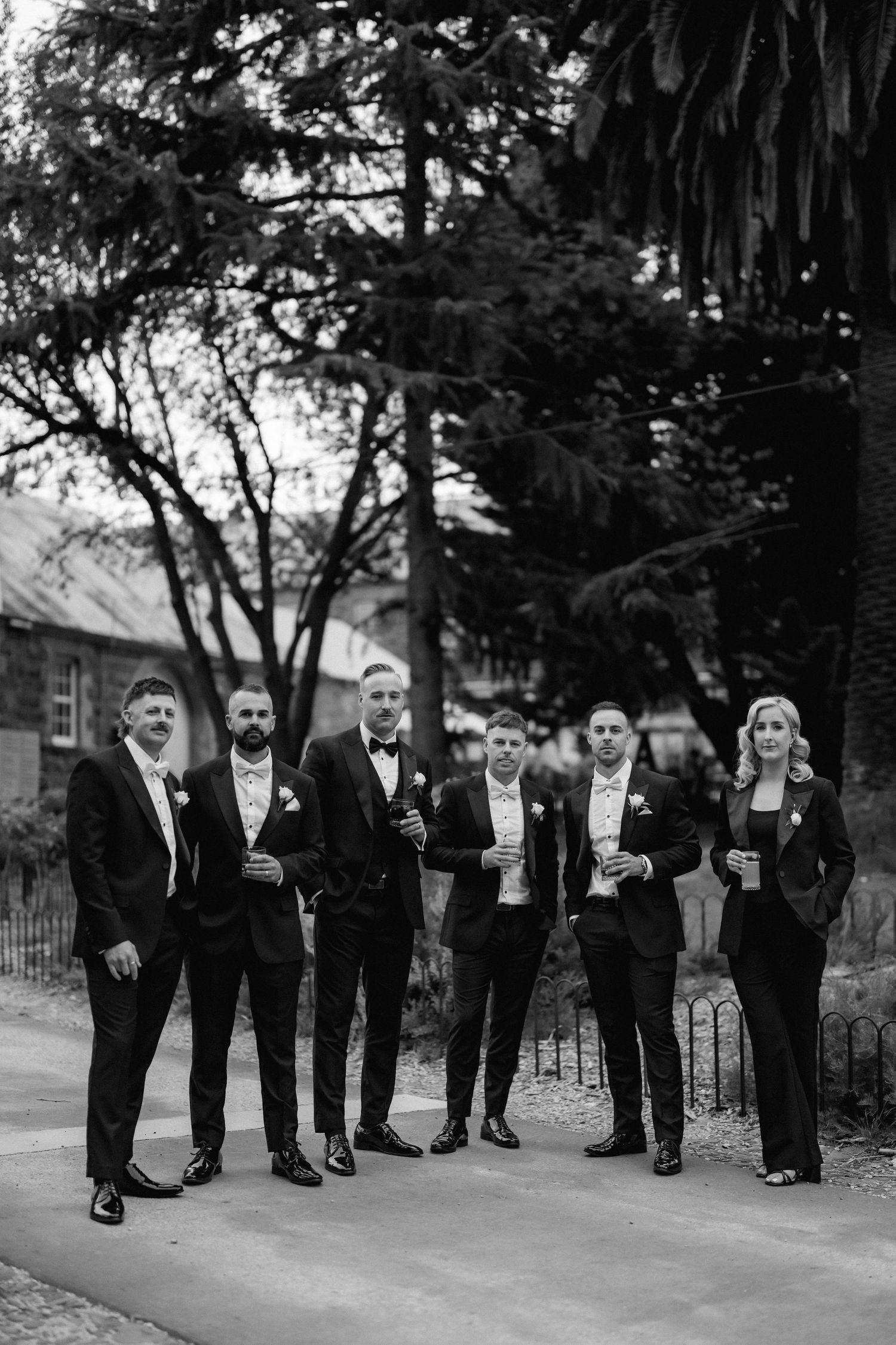 Wedding party in black formal attire poses together on outdoor path while holding champagne glasses.