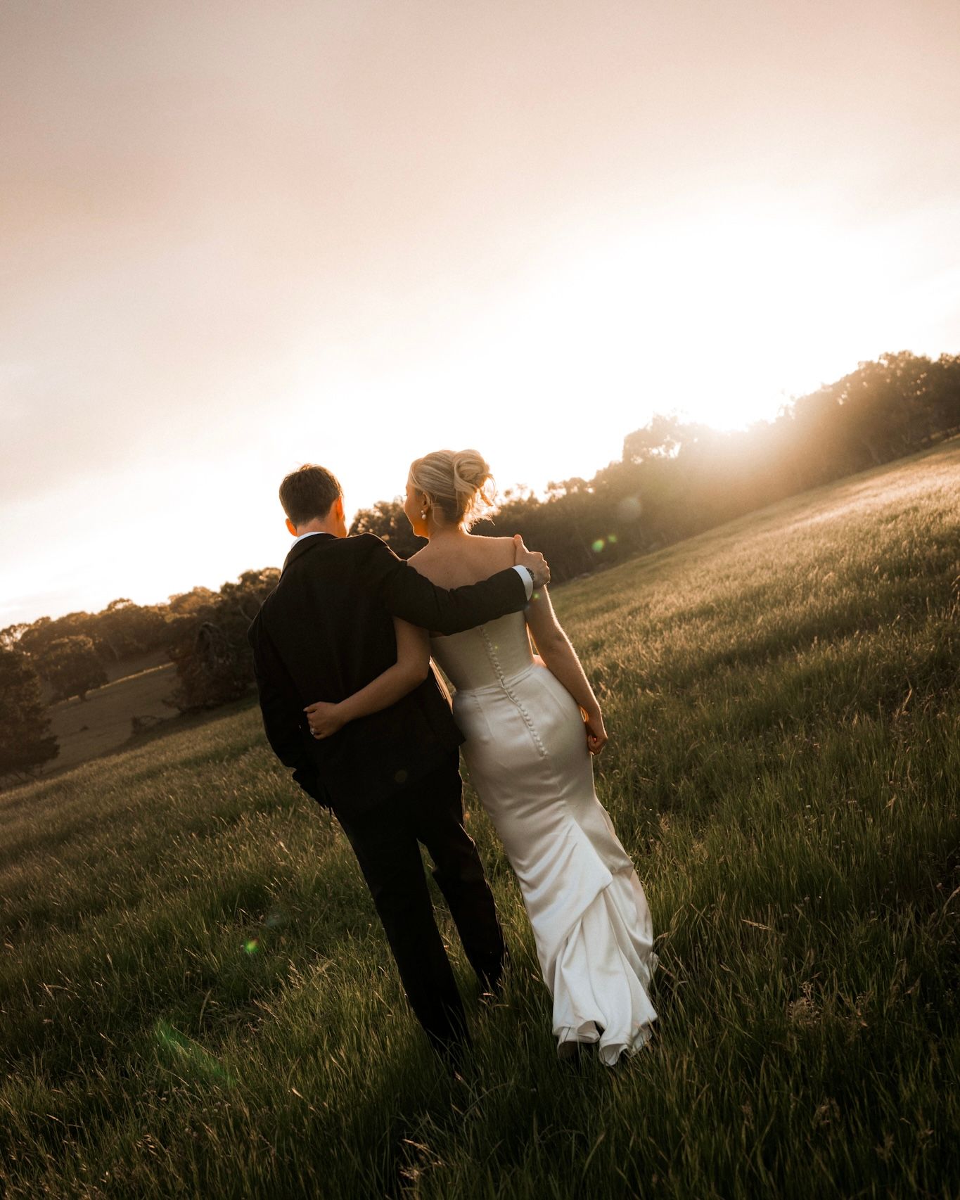 A couple in wedding attire dances together in a sunlit meadow at golden hour.