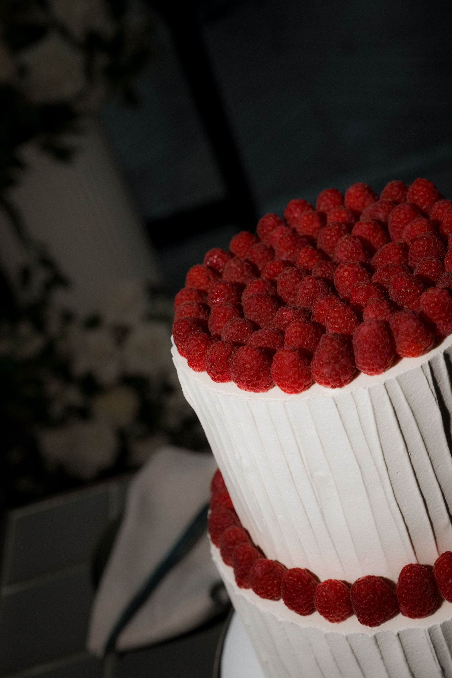 A white layered cake decorated with fresh red raspberries arranged in rows around the sides and top creates an elegant design.