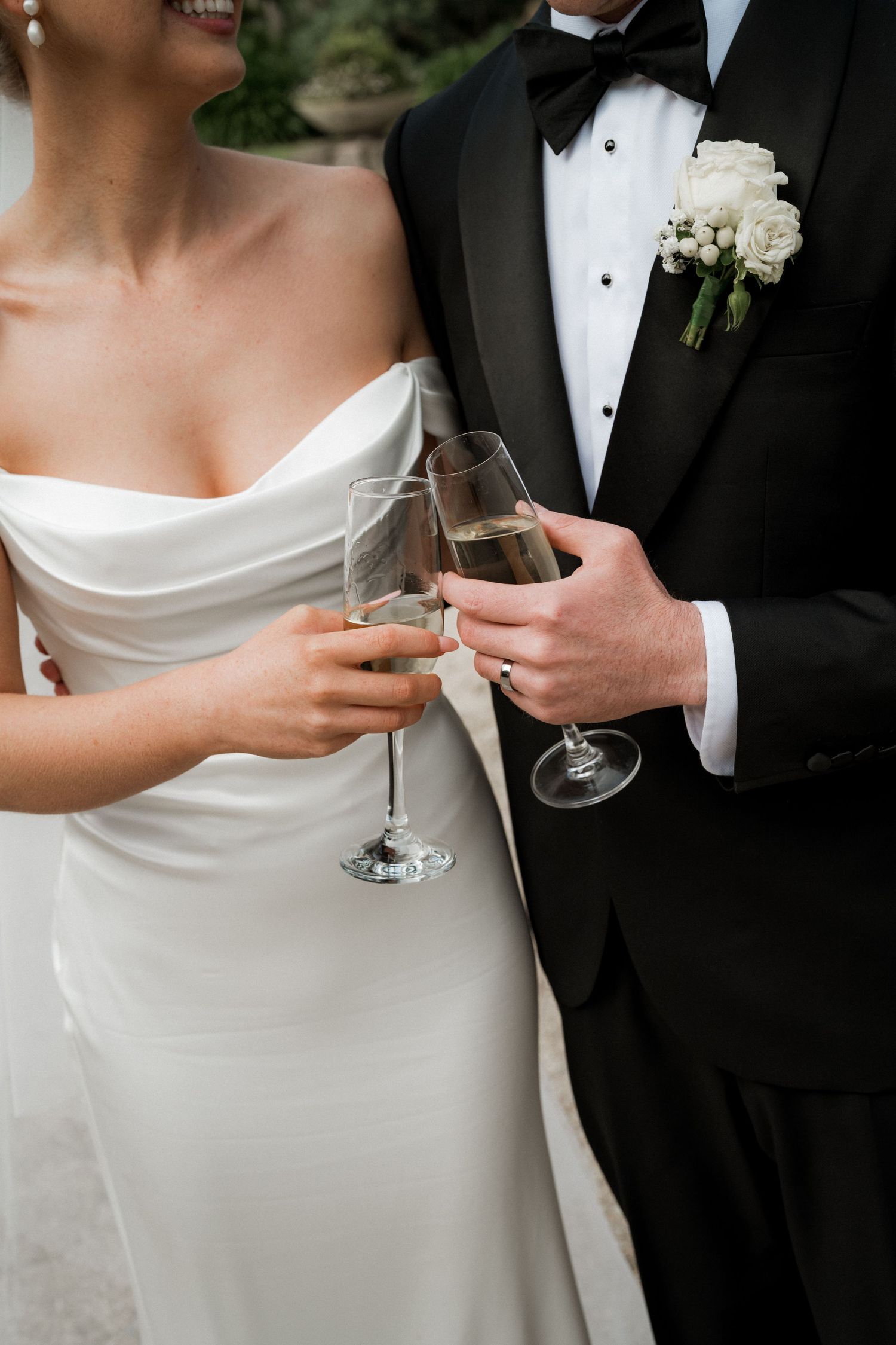 A bride in a white gown and groom in tuxedo toast with champagne glasses at their wedding celebration.