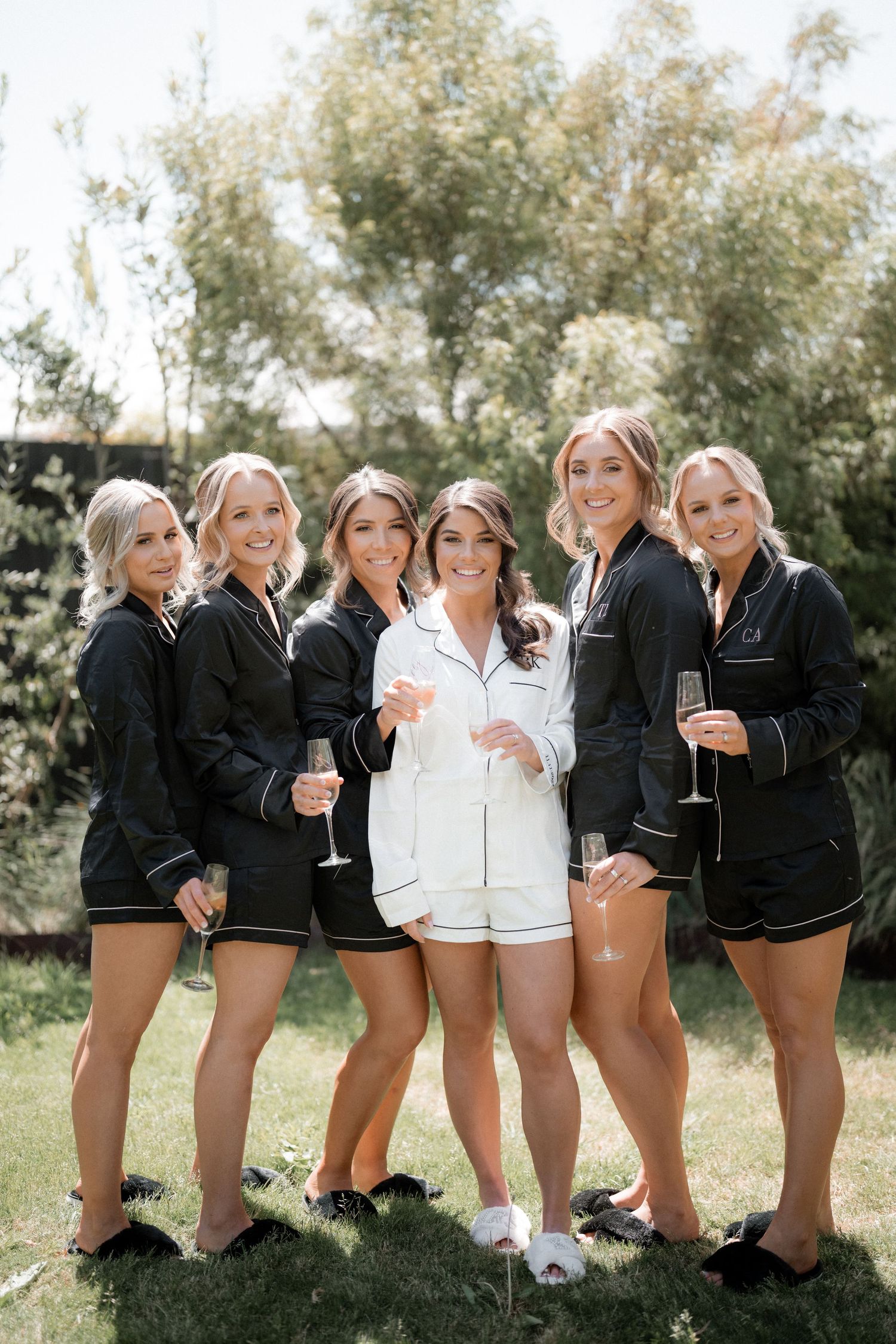 Bridesmaids in matching black jackets and white shorts pose playfully with champagne glasses outdoors.