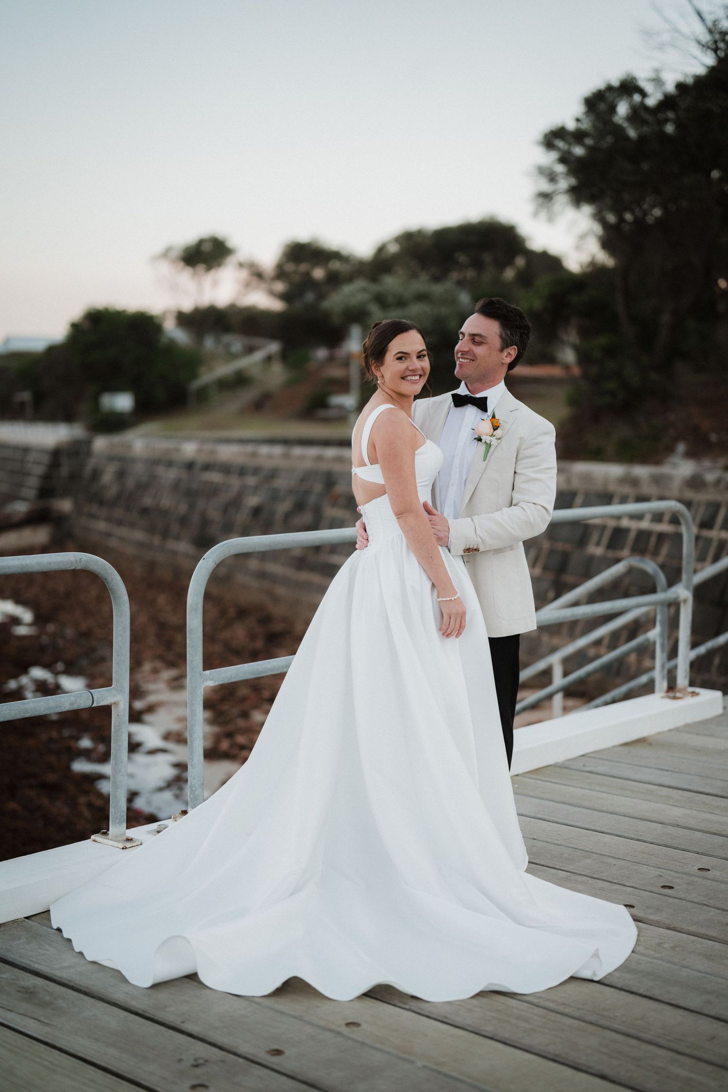 Couple in wedding attire poses together on a wooden dock at dusk.