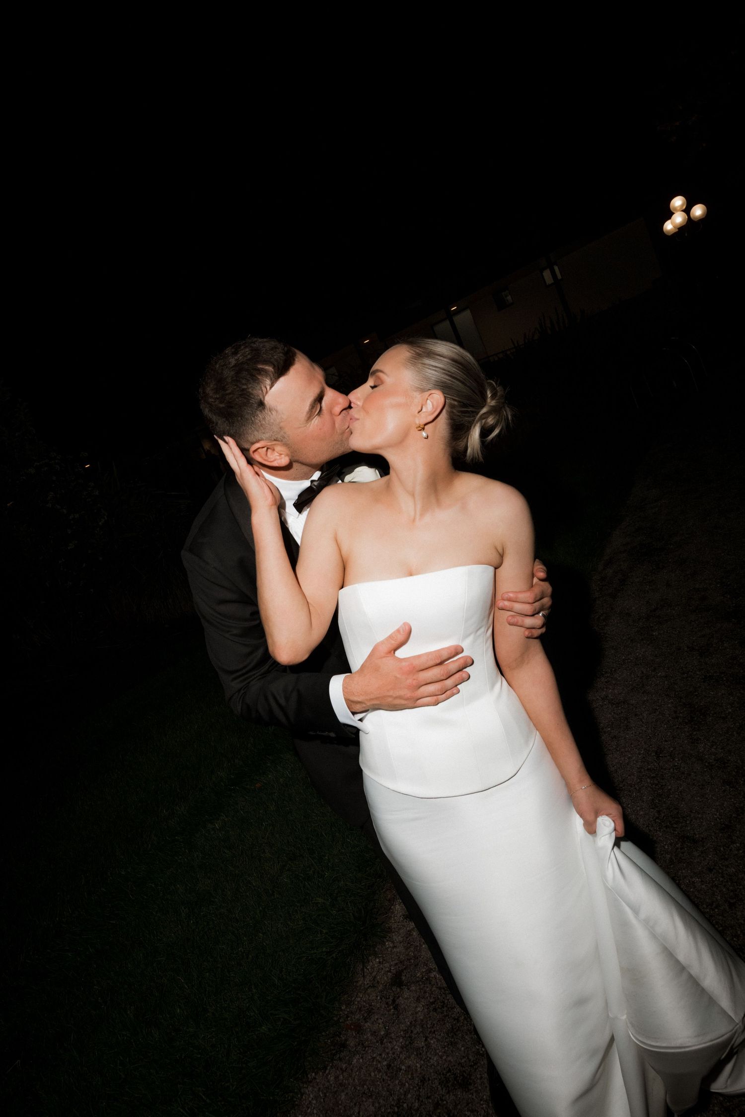 Groom kisses bride tenderly outdoors at night, bride wearing strapless white wedding gown.