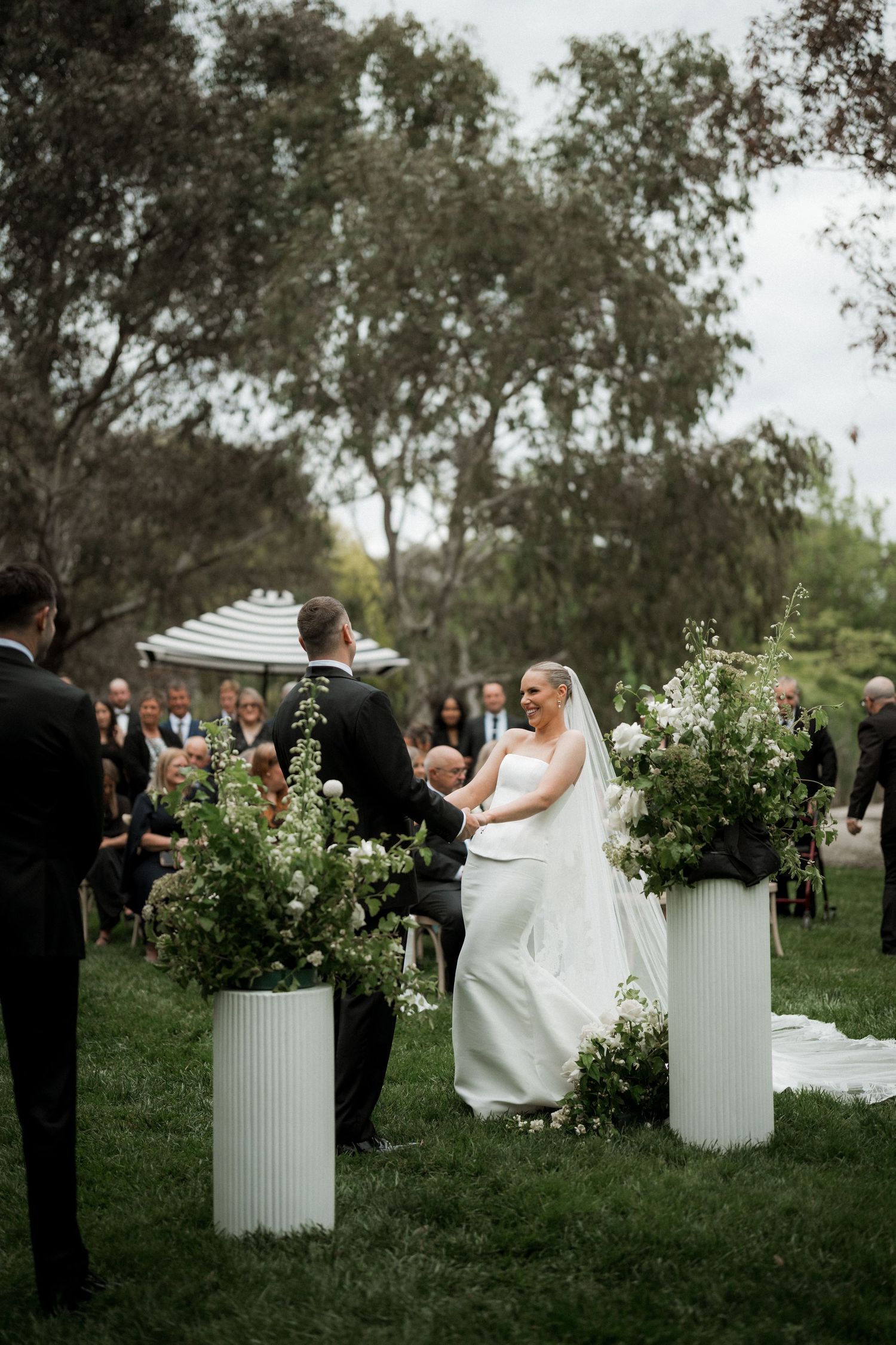 Bride and groom exchange vows at outdoor wedding ceremony with white floral pillar arrangements.