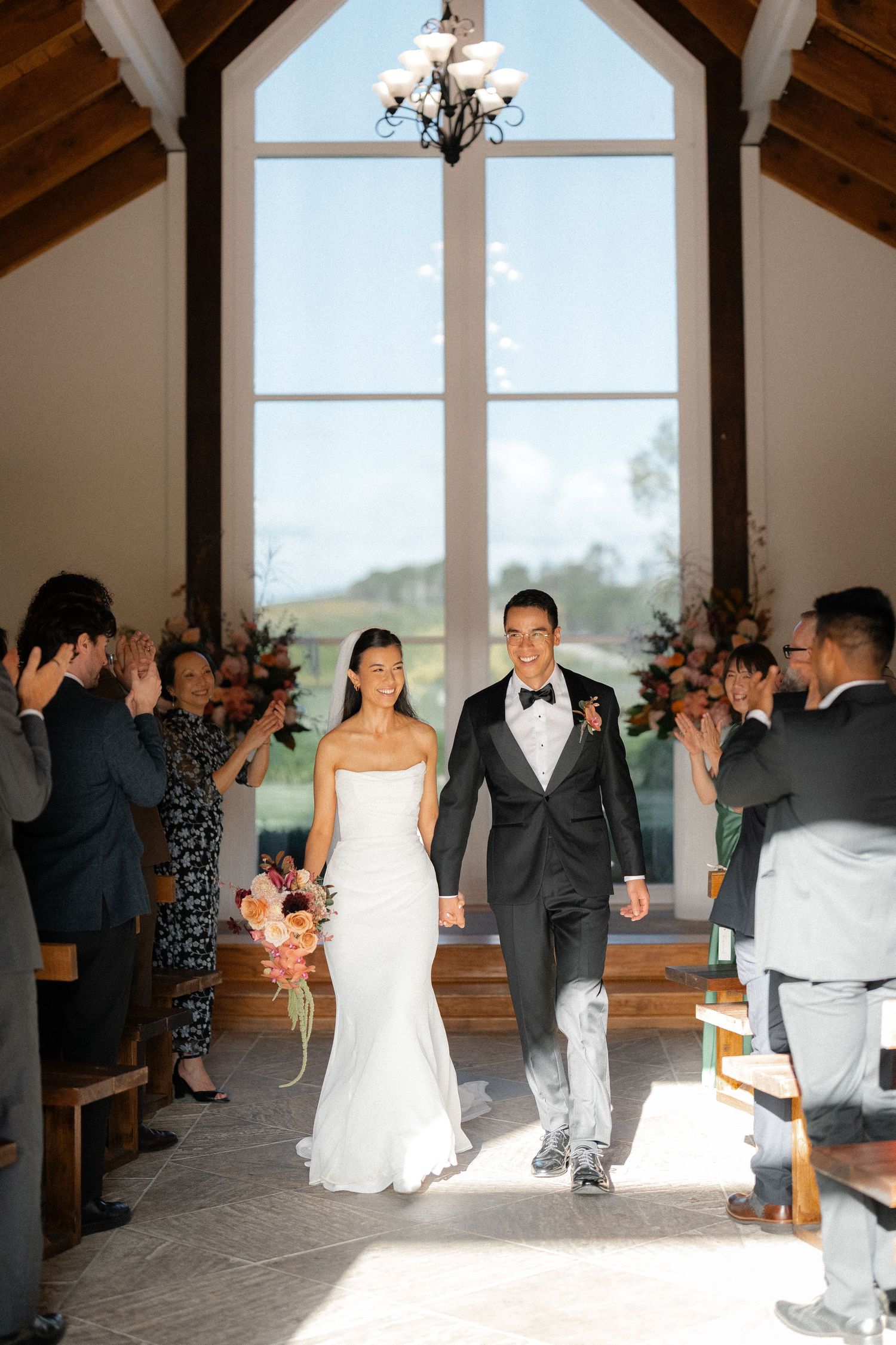 Newlyweds walk down chapel aisle as guests applaud after wedding ceremony.