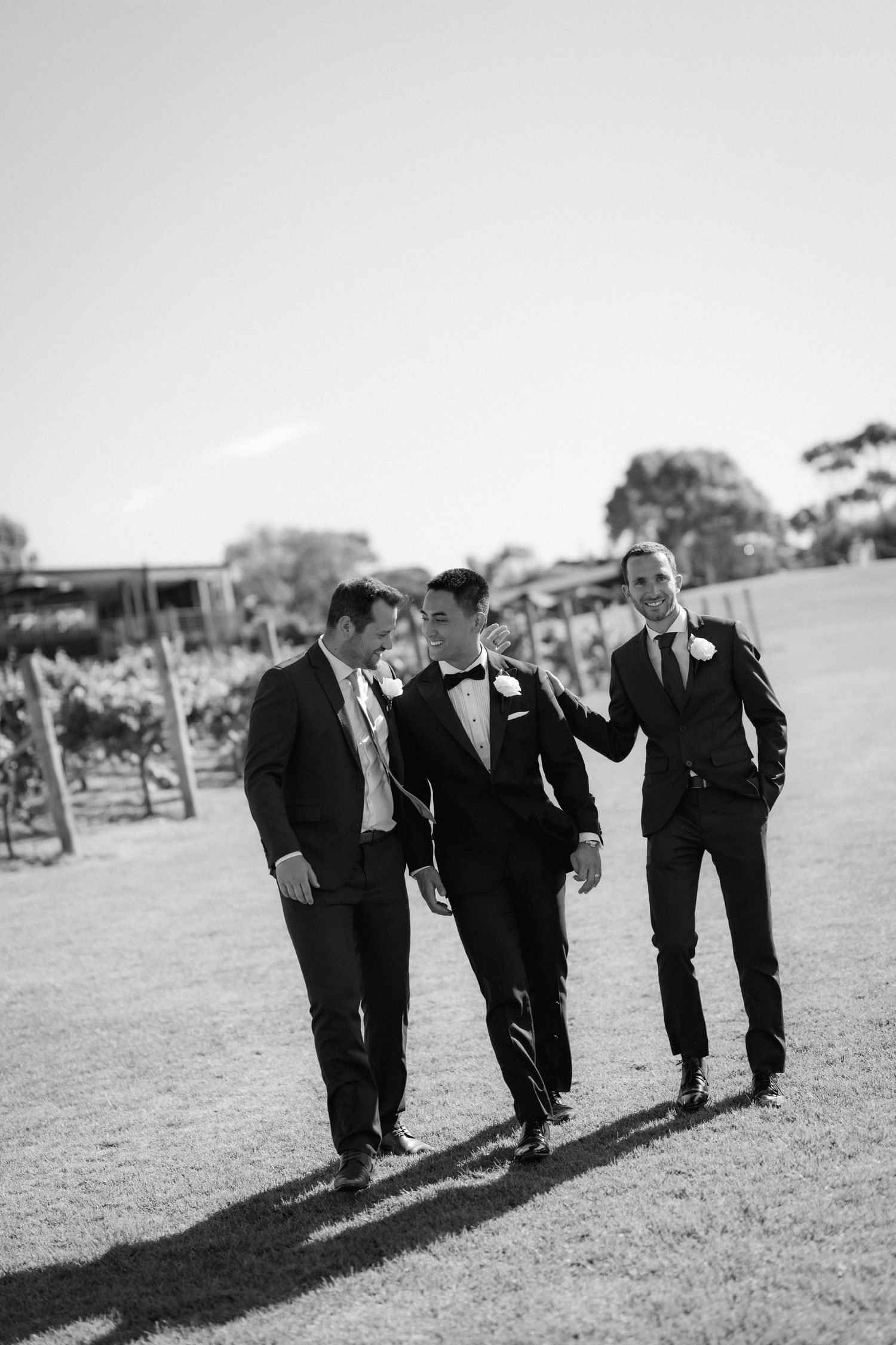 Three groomsmen walking and laughing together on grass in a casual candid black and white photo.