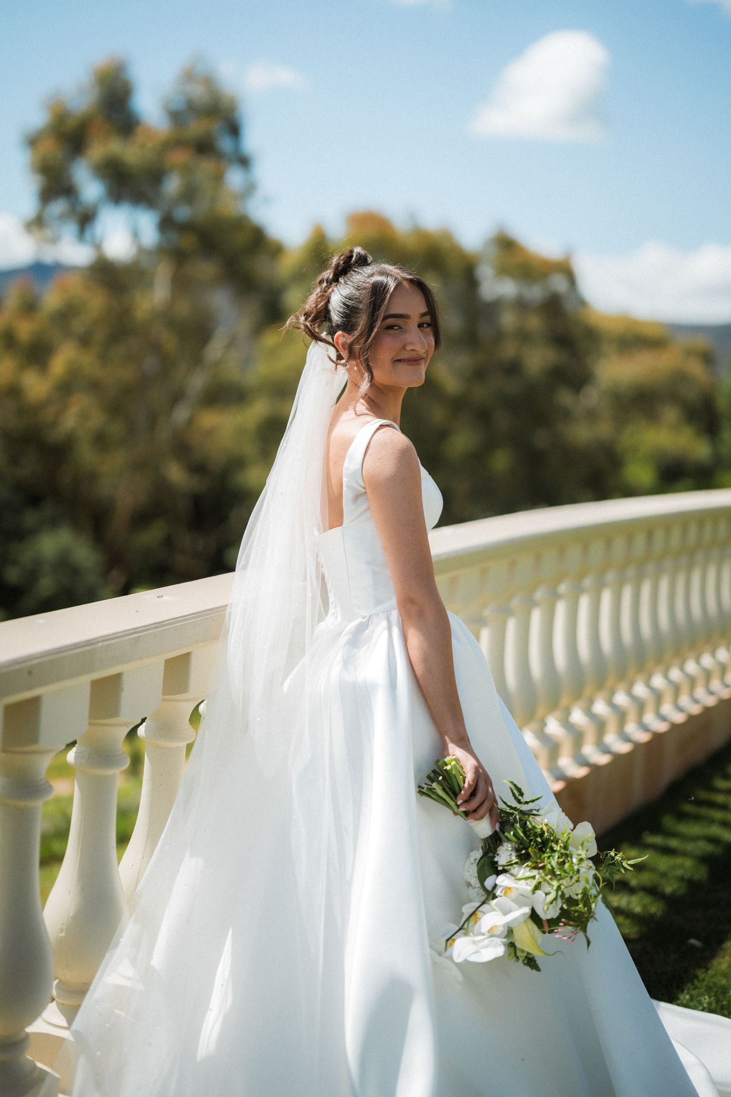 Smiling bride in a white gown and long veil holds a bouquet of white flowers on a balcony with trees and blue sky behind her.