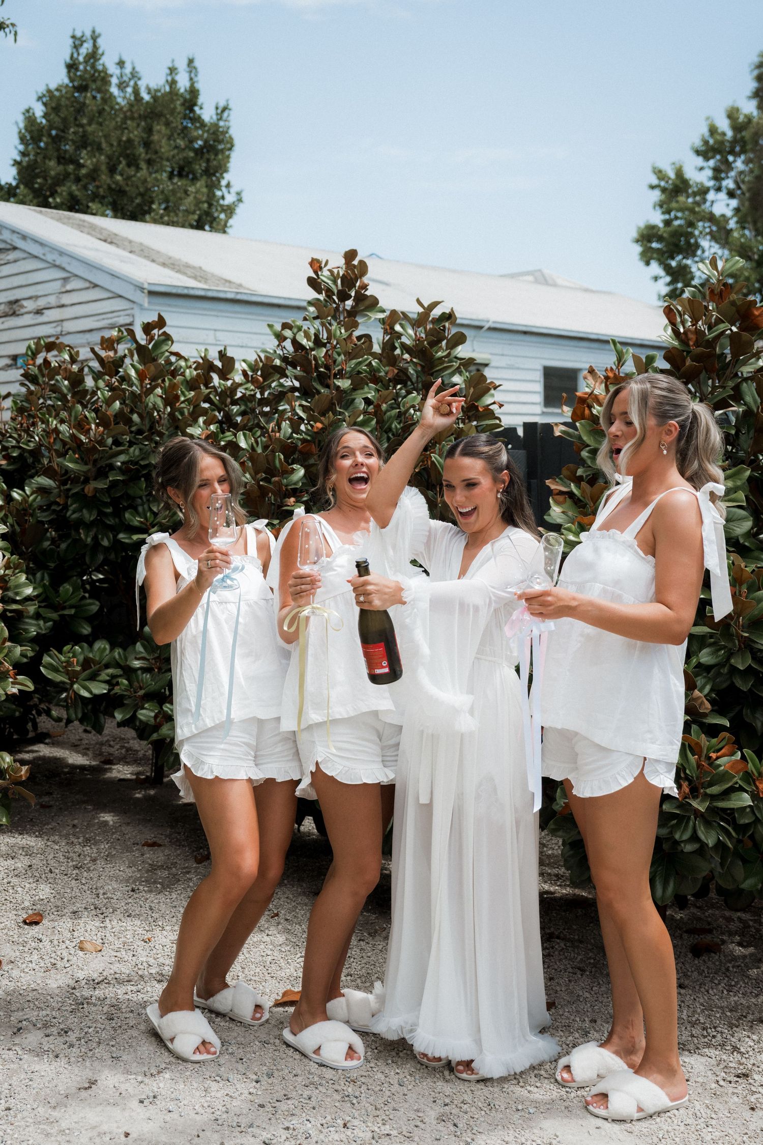 Bridesmaids in white robes celebrating with drinks outdoors by magnolia trees.