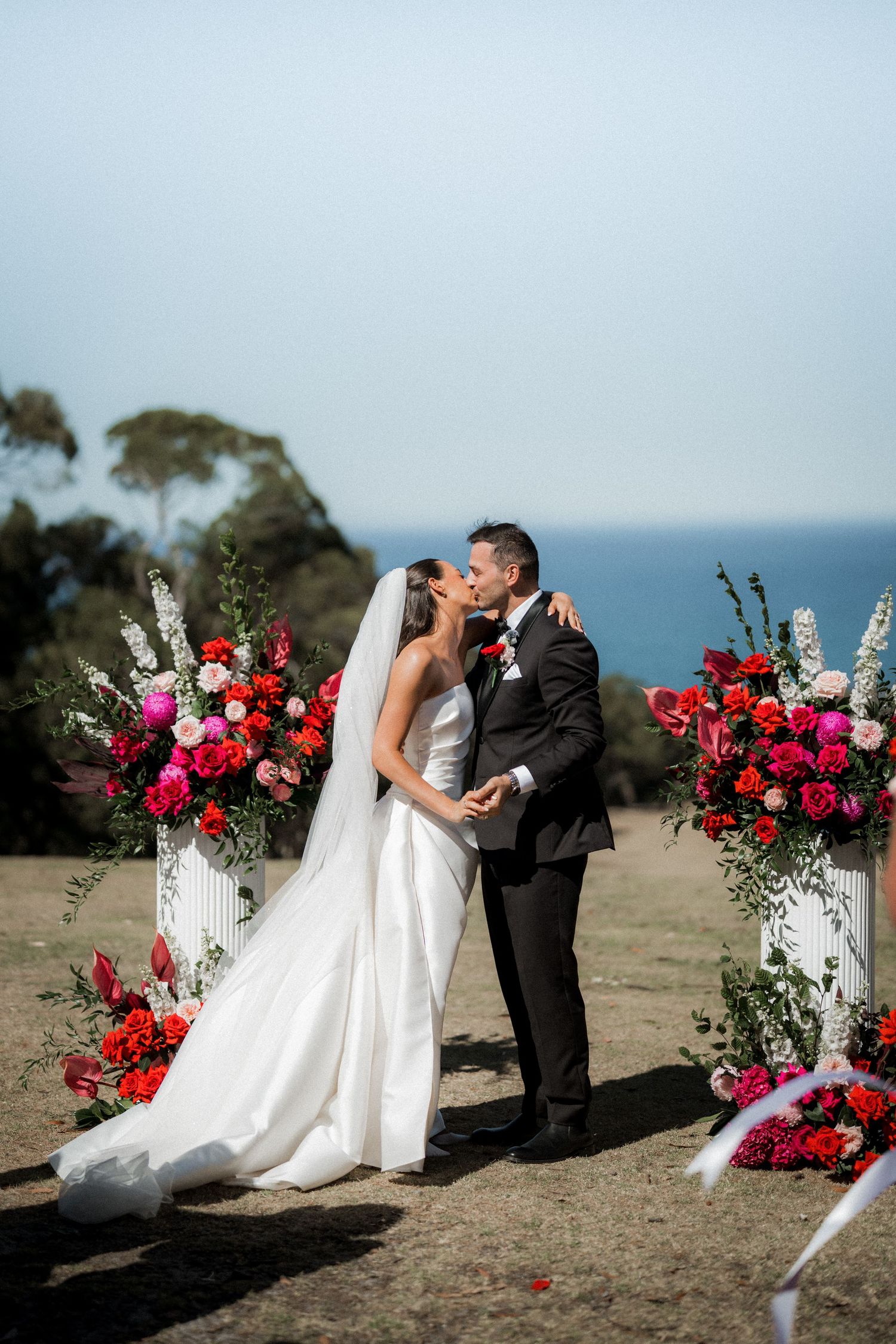Couple shares romantic moment by floral arch with red roses against ocean backdrop.