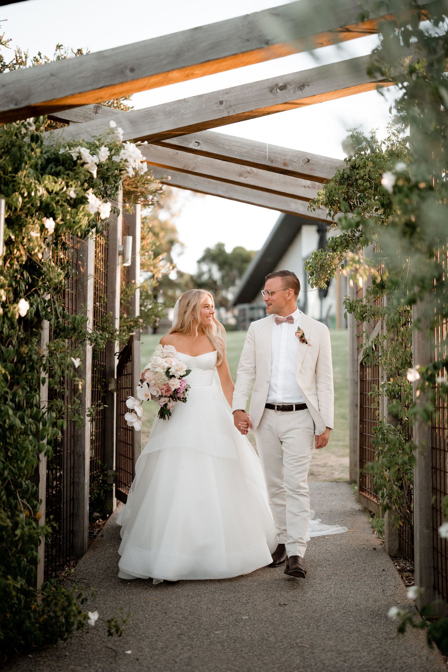 A couple walks through a garden pergola decorated with white flowers and greenery for their wedding celebration.