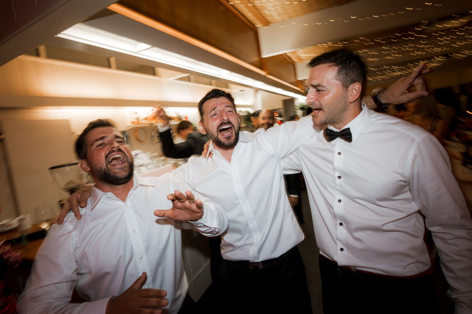 Groomsmen in white shirts celebrating and laughing together at an indoor wedding reception.