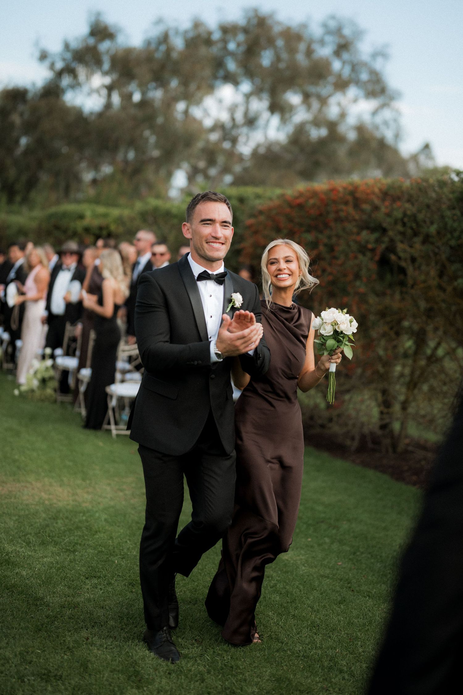 Wedding party members in formal attire walking and laughing on a green lawn.