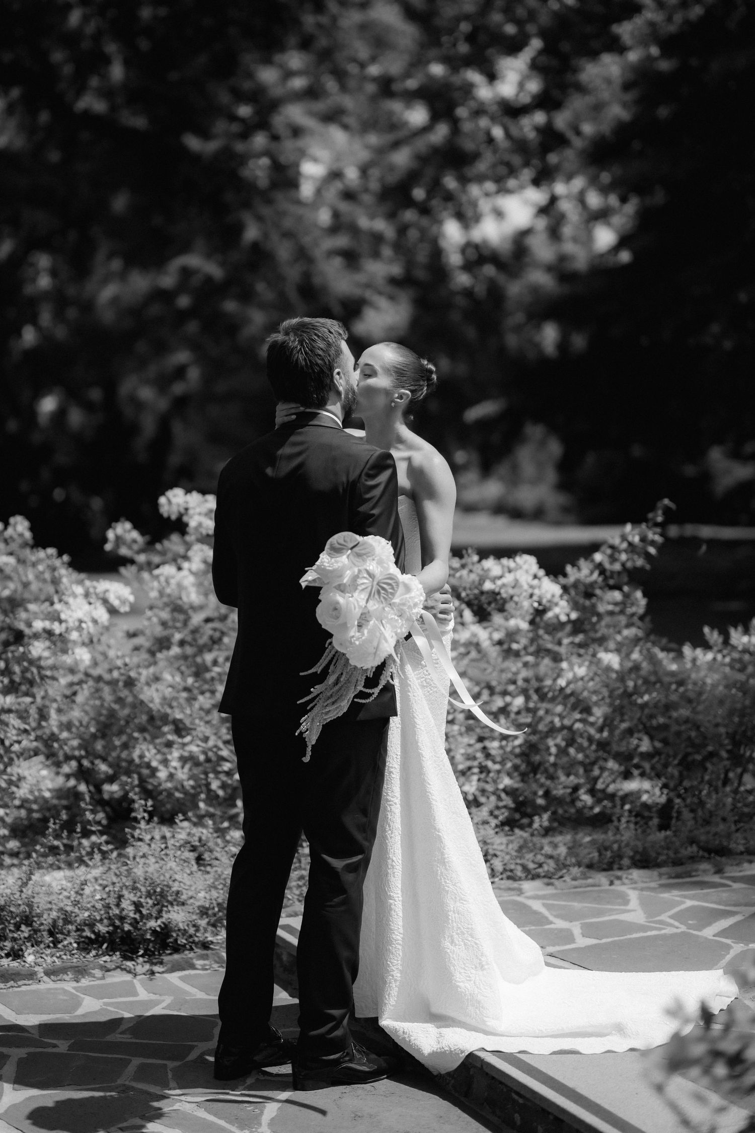 Black and white romantic photo of bride and groom sharing a kiss in a lush garden on their wedding day.