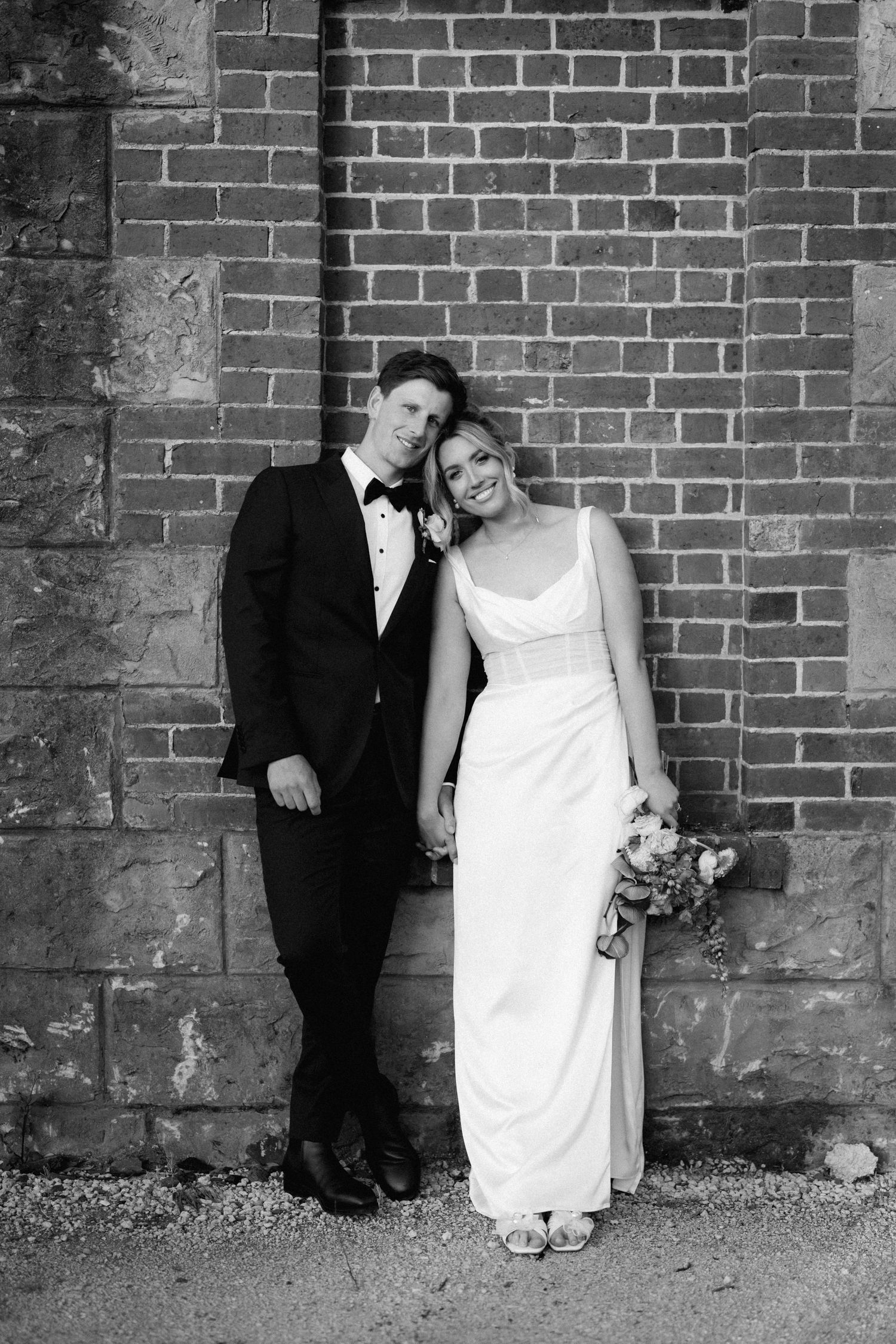 Newlyweds pose against a brick wall in formal attire.