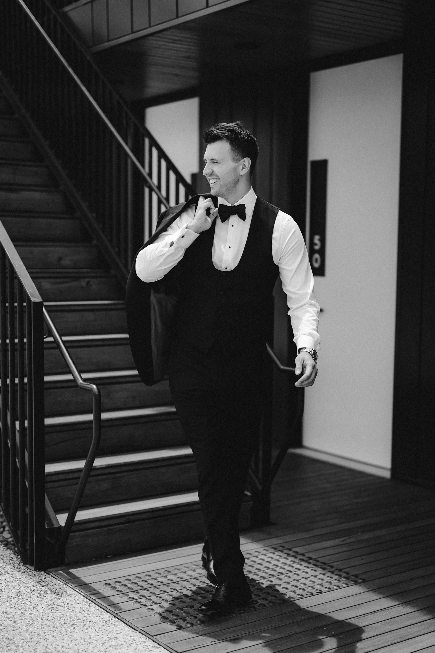 Black and white photo of someone in formal attire with a bowtie and vest standing on wooden stairs in contemplative pose.
