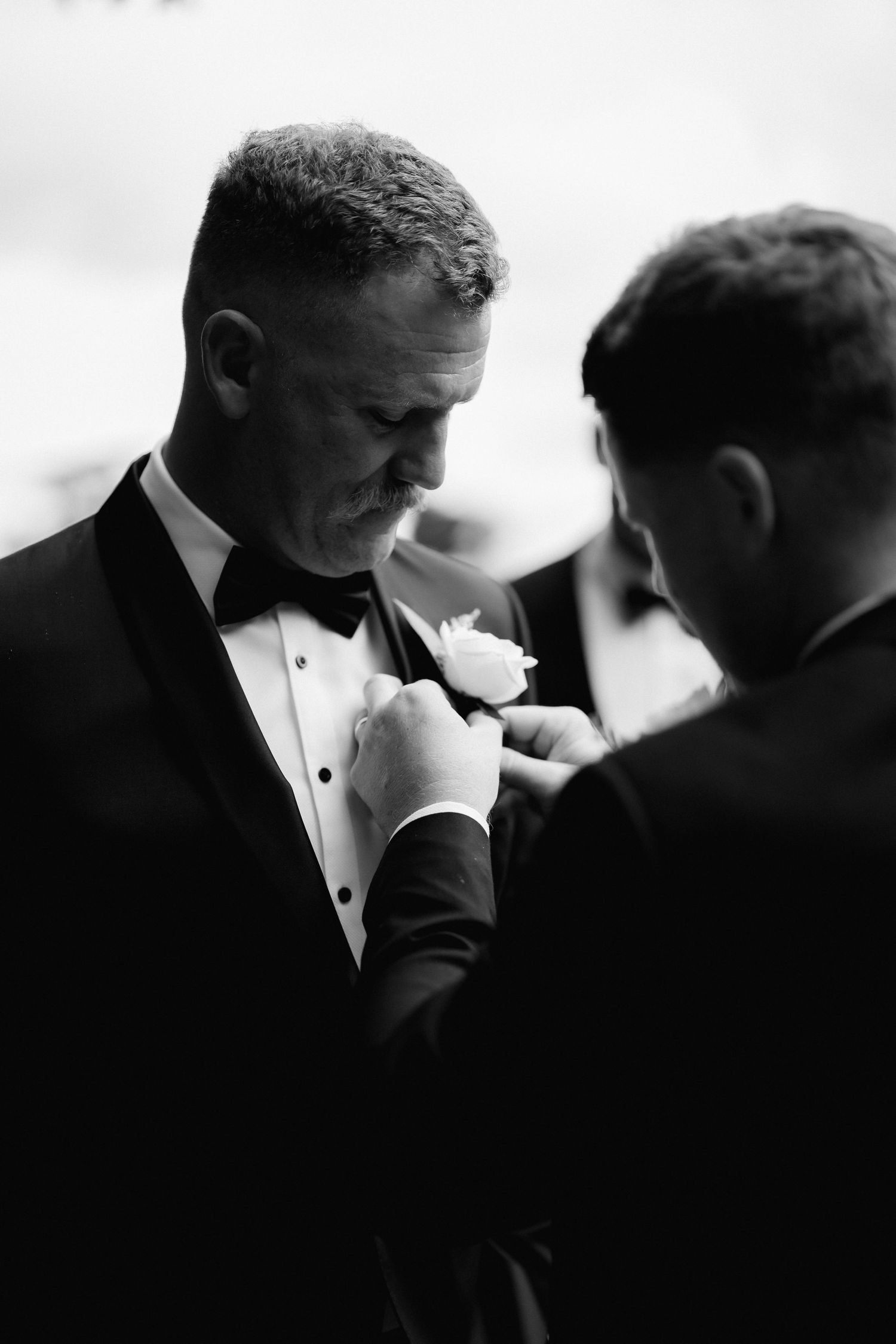 Black and white photo showing someone adjusting a boutonniere on a man's tuxedo lapel during a formal event.