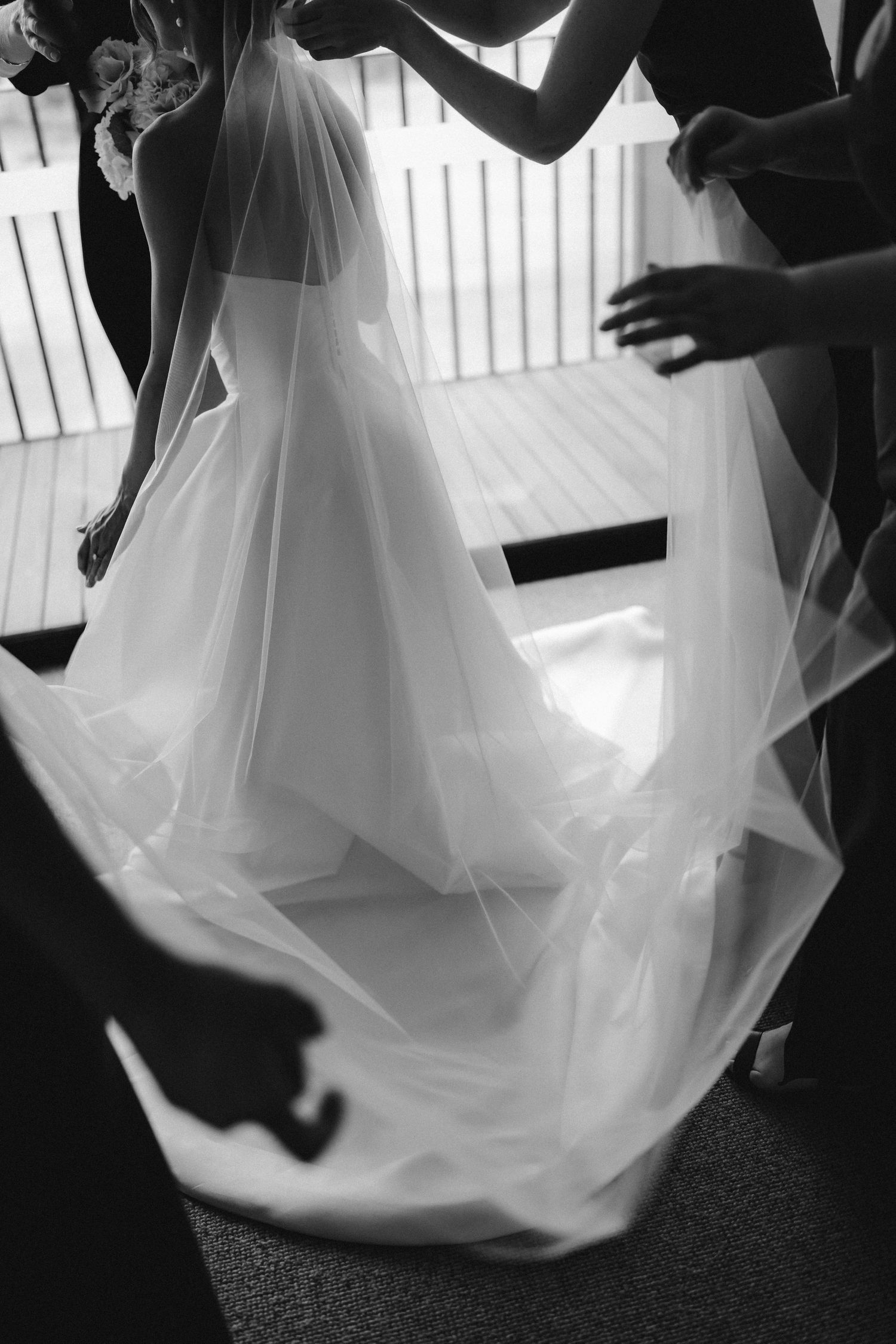 Black and white photo of ethereal wedding veil being arranged on floor with hands visible in silhouette.