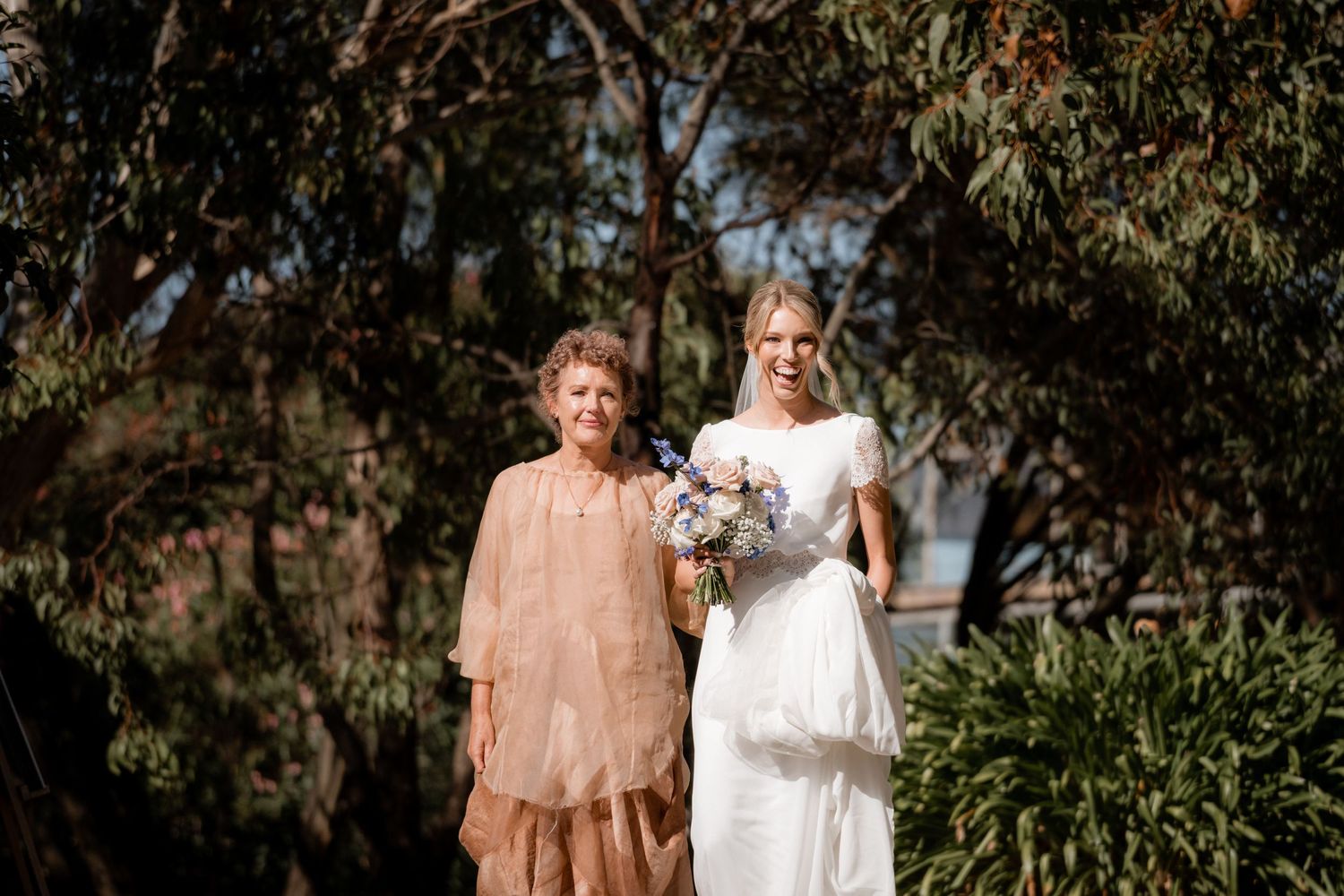 Two figures stand together outdoors in formal attire against a backdrop of eucalyptus trees on a sunny day.